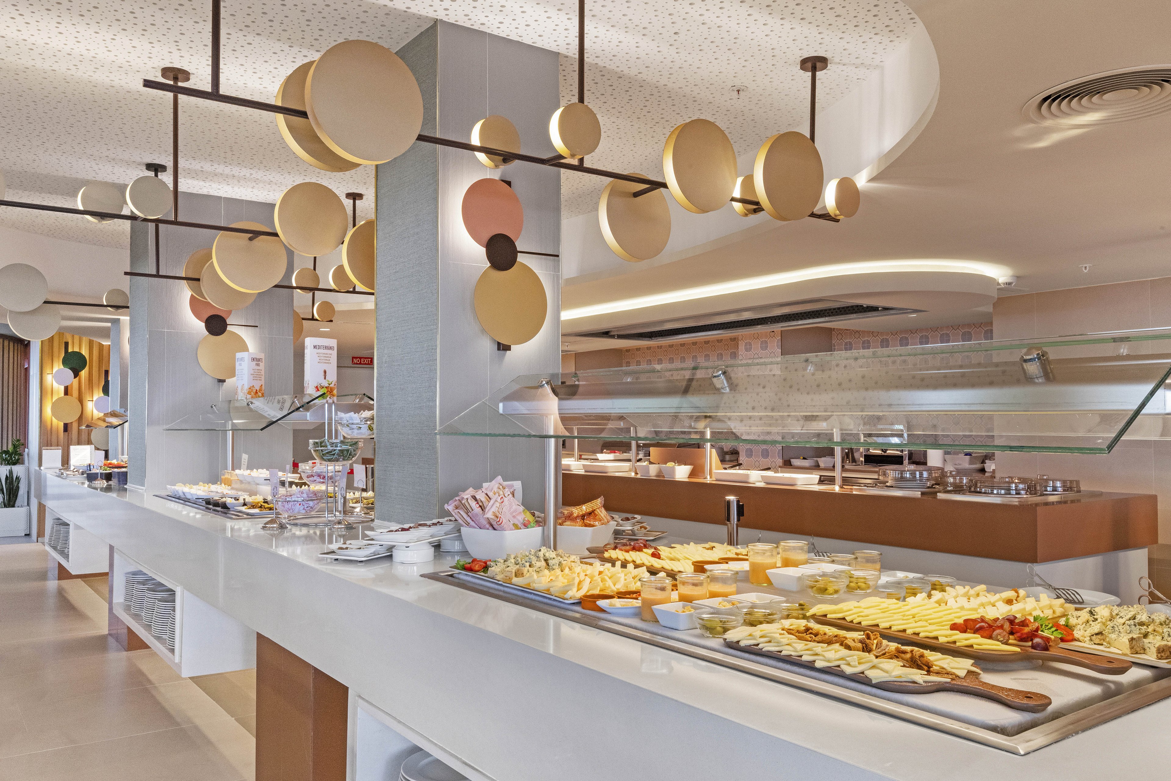 A spacious, contemporary buffet area displays a variety of breakfast foods, including cheeses, pastries, and fruits. The setting features sleek white countertops, geometric lighting fixtures, and a clean, inviting atmosphere. The buffet is well-organized with plates and utensils ready for guests. No visible people are present, emphasizing the food presentation. The overall mood is bright and welcoming, with a neutral color palette.