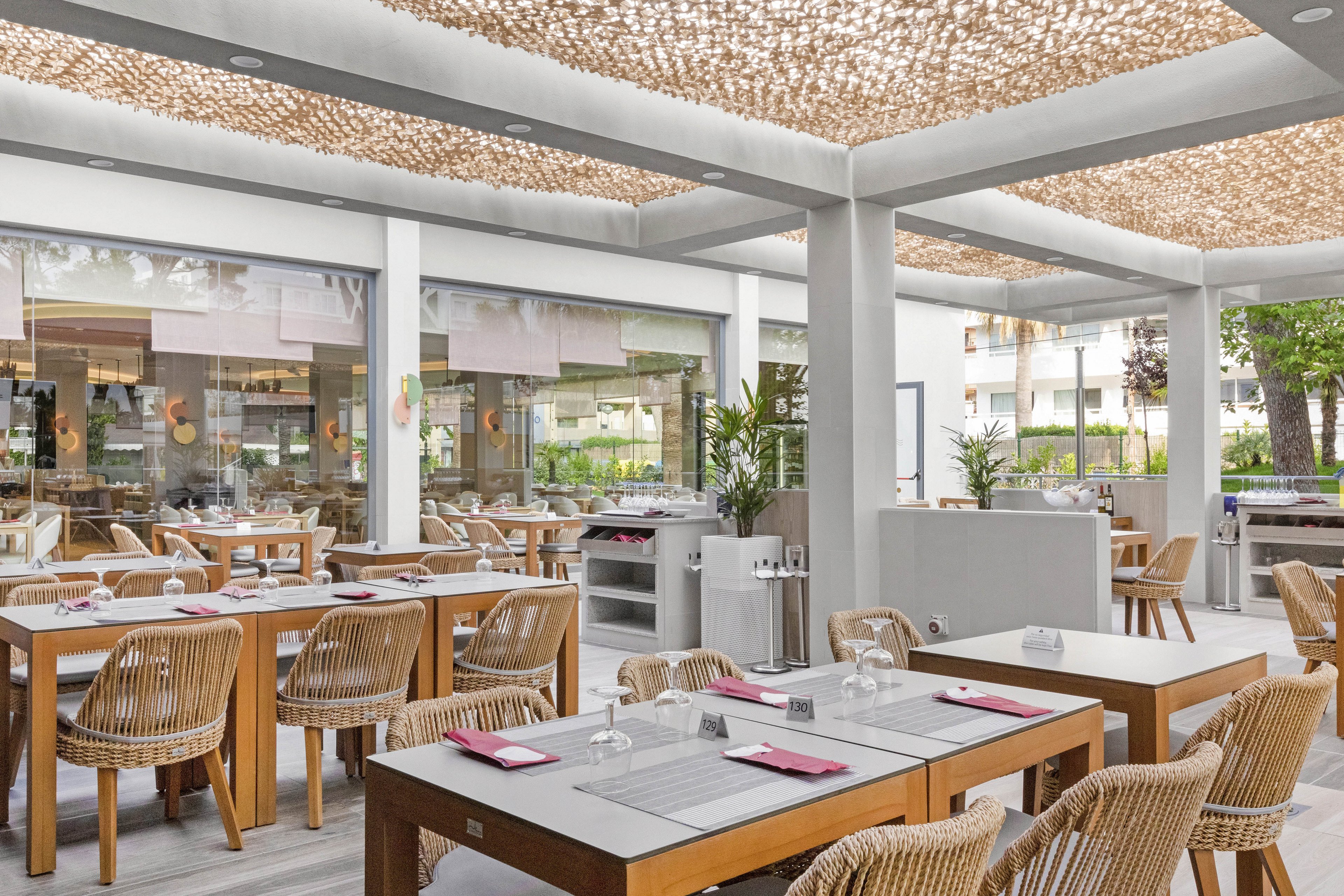A contemporary outdoor restaurant patio features wooden tables and wicker chairs arranged neatly under a stylish canopy. The setting is bright and airy, with natural light filtering through the patterned ceiling and lush greenery visible outside. Table settings include glassware, napkins, and cutlery, creating a welcoming dining atmosphere. The overall palette is neutral with accents of natural wood and greenery, evoking a relaxed and inviting mood.