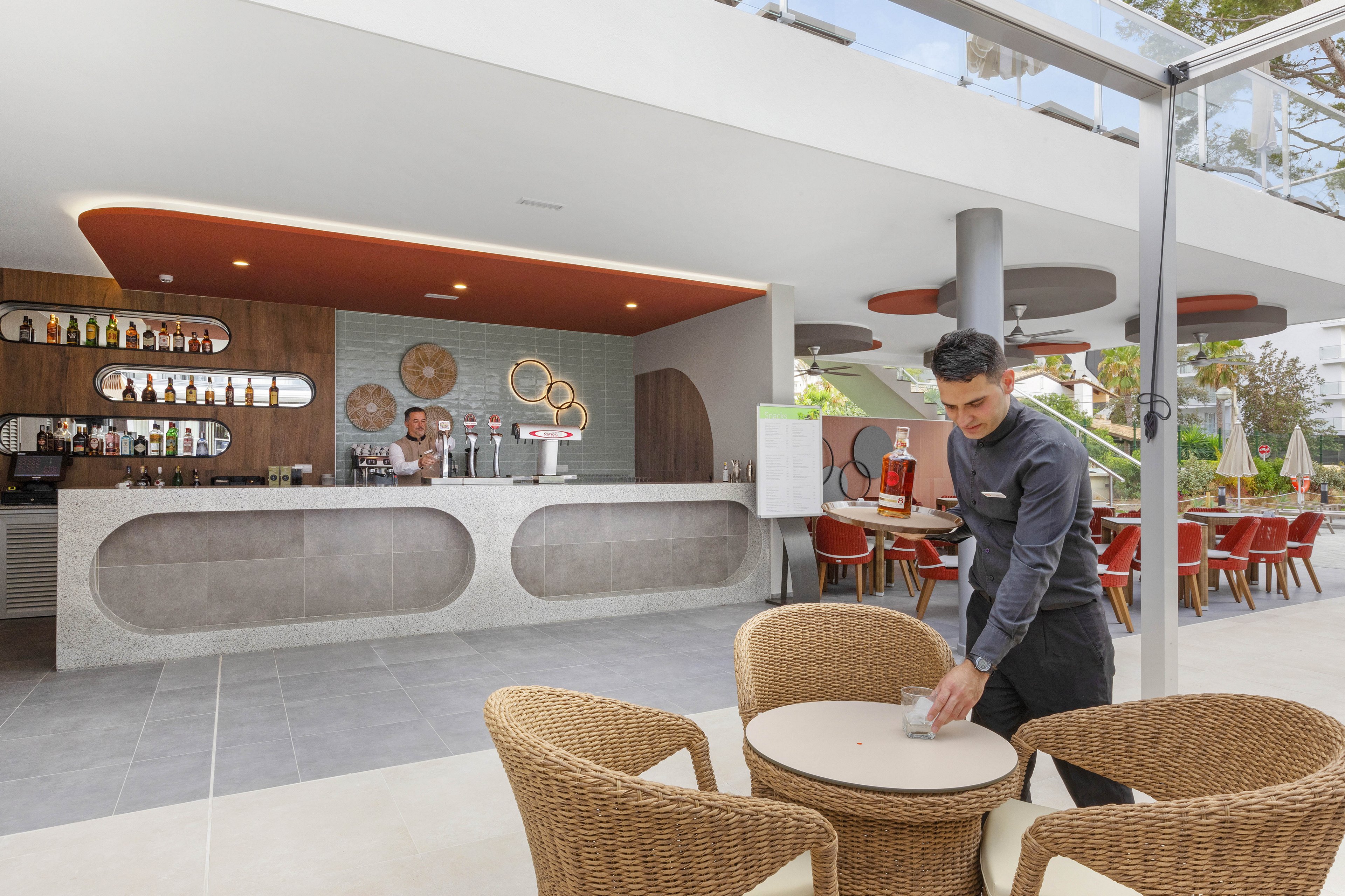 A contemporary outdoor bar features a sleek counter and a well-stocked back wall. A male server is clearing a small round table with wicker chairs in the foreground, while two bartenders work behind the counter. The setting is bright and airy, with red and neutral tones, and visible glassware and bottles add detail to the scene.