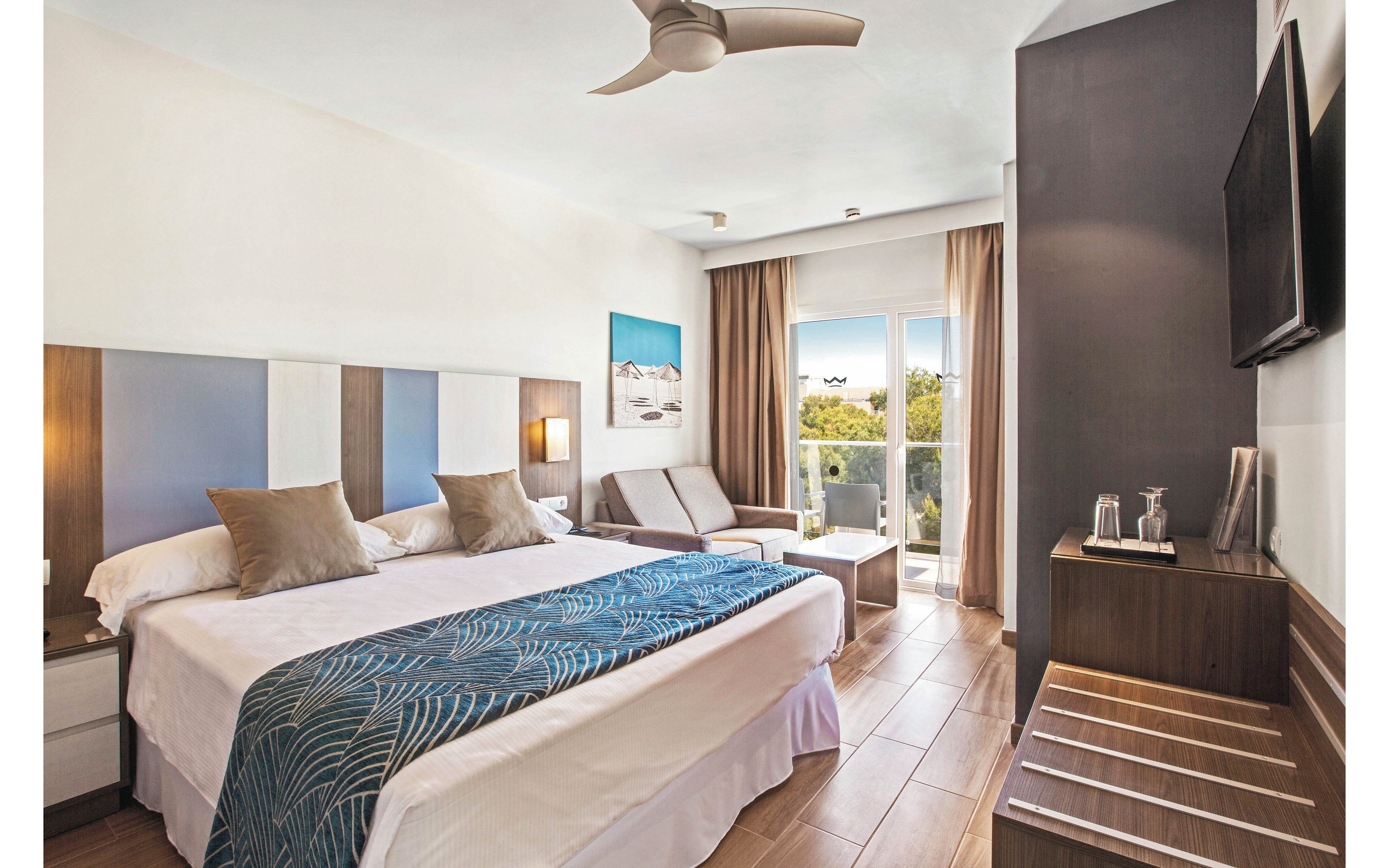 A contemporary hotel room features a large king-size bed with decorative pillows and a patterned runner. The space includes a small sitting area, a wall-mounted television, and a balcony with glass doors letting in natural light. The room is styled in neutral tones with wood accents, and amenities such as a kettle and cups are visible on a side table. The overall mood is calm and inviting, ideal for relaxation or business travel.