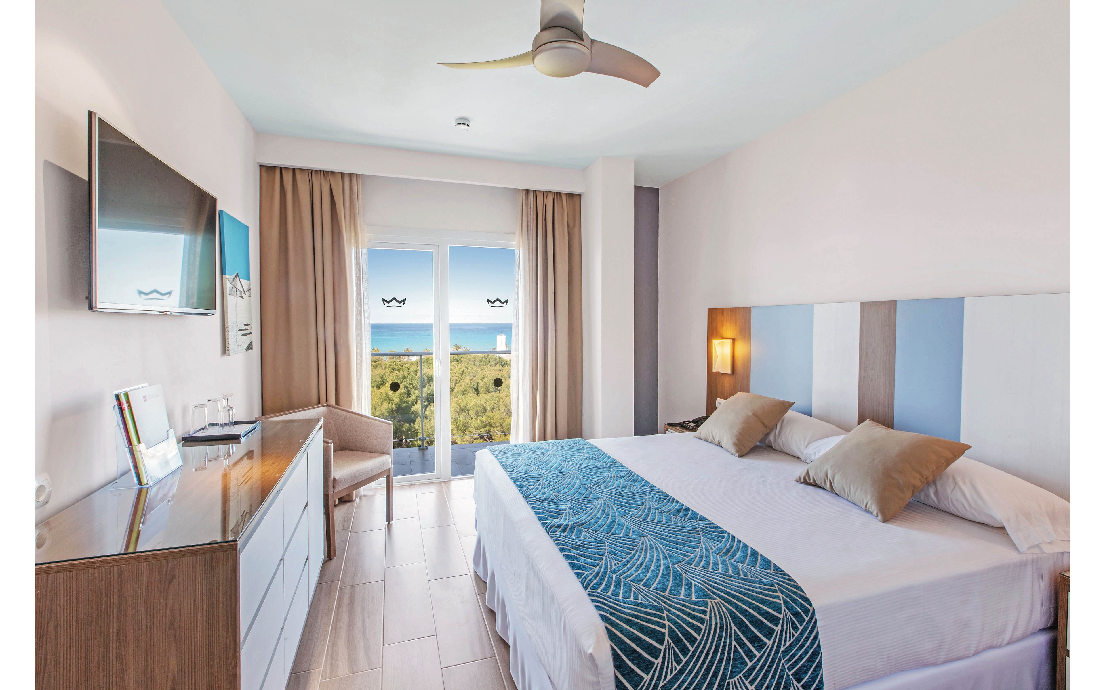 A bright, contemporary hotel room features a large double bed with blue and beige accents, a wall-mounted television, and a dresser. The room opens to a balcony with glass doors, offering a scenic view of the ocean and greenery. Natural light fills the space, creating a calm and inviting atmosphere, with minimalistic decor and neutral tones.