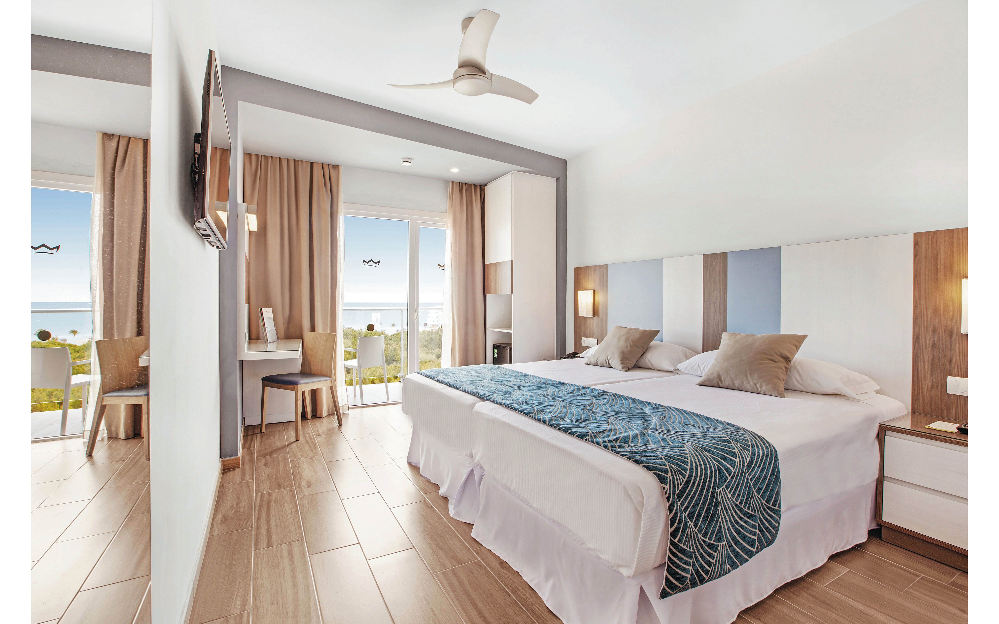 A spacious hotel room features a large bed with patterned blue runner and neutral pillows. The room is bright, with wood flooring, minimalist decor, and a ceiling fan. Floor-to-ceiling windows open to a balcony overlooking the ocean, with two chairs and a desk visible. The palette is light and airy, emphasizing relaxation and comfort.