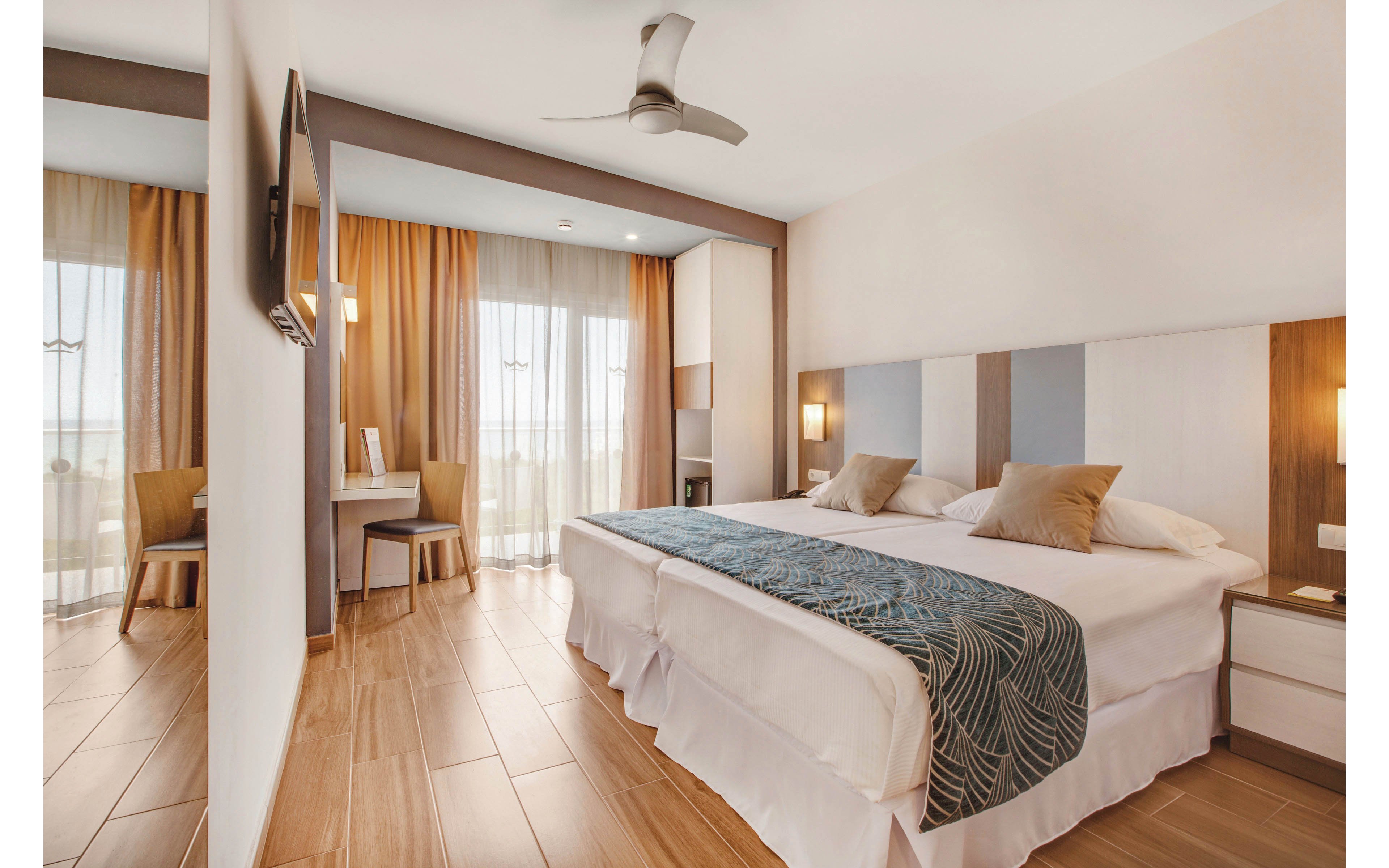 A spacious hotel room features a large king-sized bed with a decorative runner and two pillows. The room is styled in neutral tones with wood flooring, contemporary furniture, and a ceiling fan. Natural light streams in through floor-to-ceiling windows with sheer curtains, leading to a balcony. A wall-mounted television and a small desk area are also visible, creating a comfortable and inviting atmosphere.