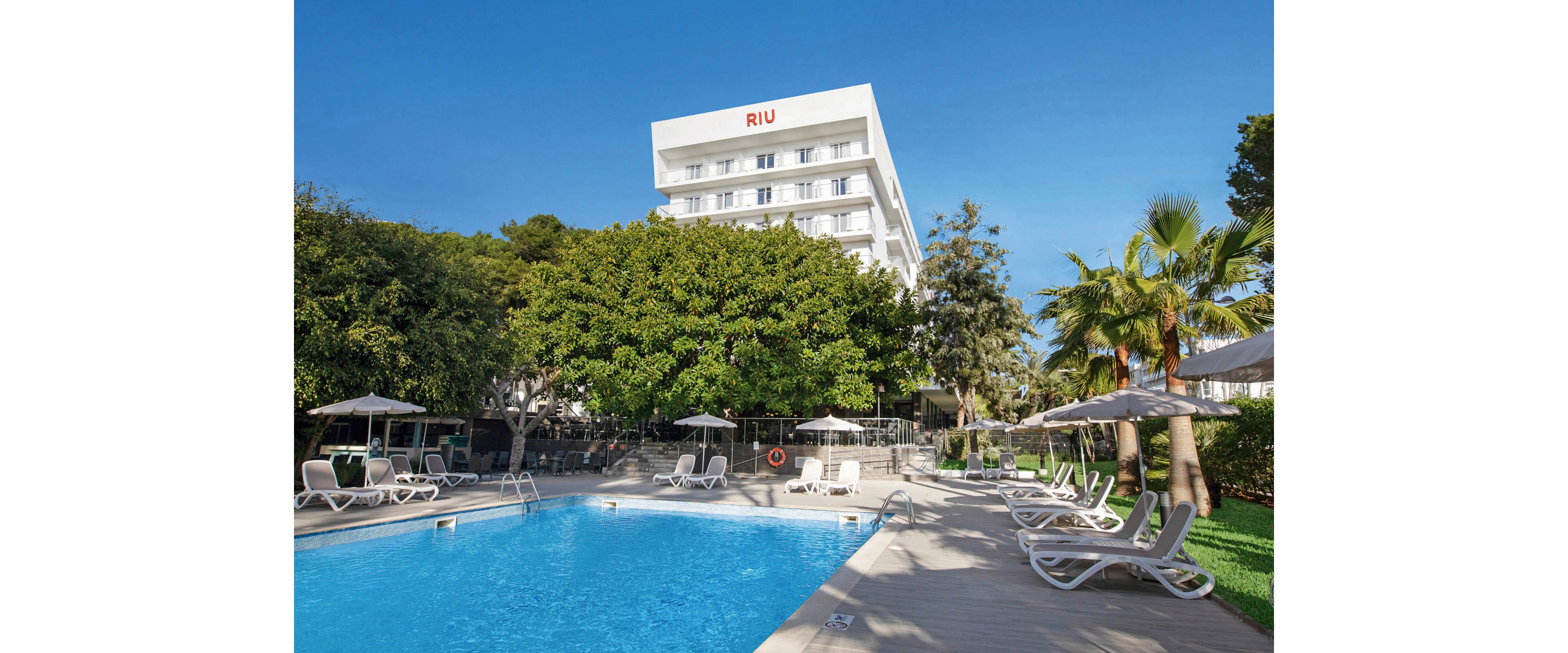 A modern RIU hotel building stands behind a large outdoor swimming pool surrounded by sun loungers and umbrellas. The setting is a sunny day with clear blue skies, lush green trees, and palm trees adding to the resort atmosphere. The pool area is clean and inviting, with a relaxed vacation mood. No people are visible in the scene, emphasizing tranquility and leisure. The word 'RIU' is clearly visible on the hotel facade.