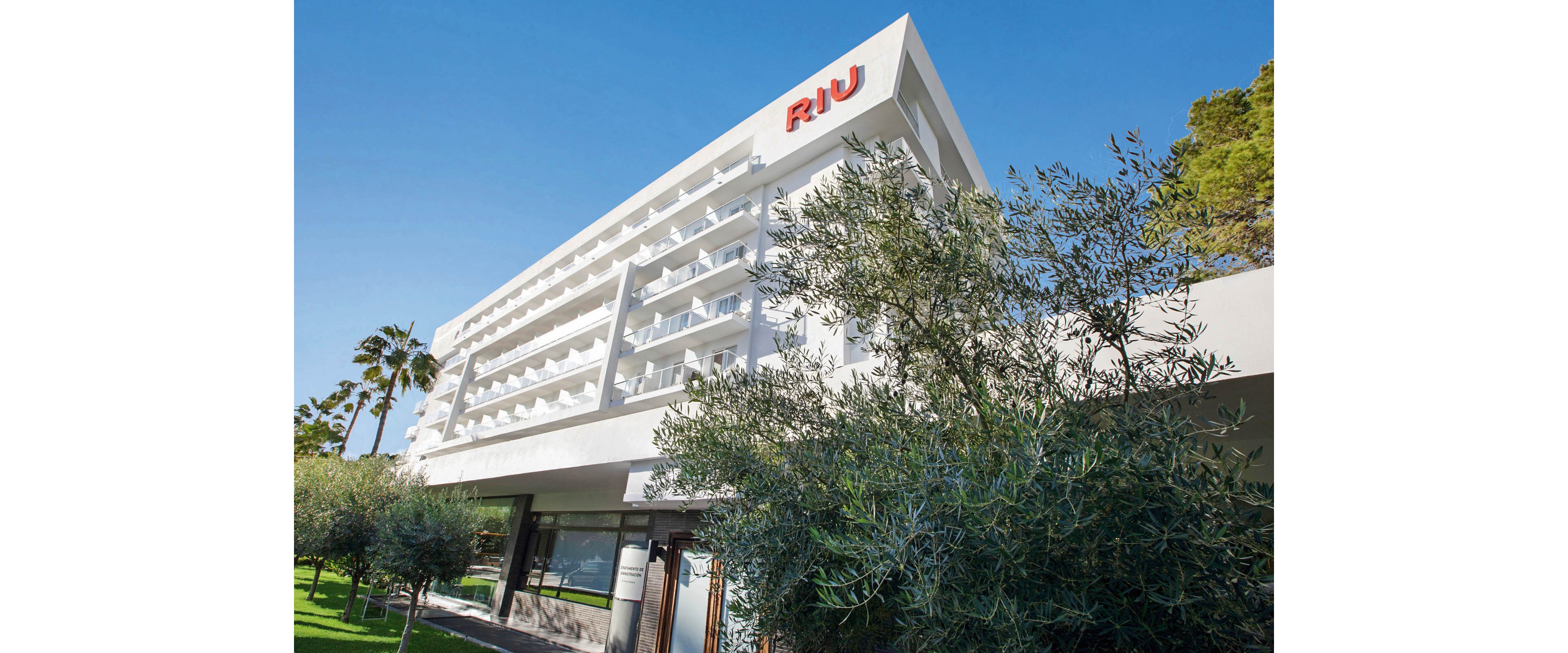 A contemporary white hotel building features the unmistakable red RIU sign on its upper facade. The structure is surrounded by lush greenery, including palm trees and shrubs, under a clear blue sky. The building's balconies and clean architectural lines create a welcoming and modern atmosphere.