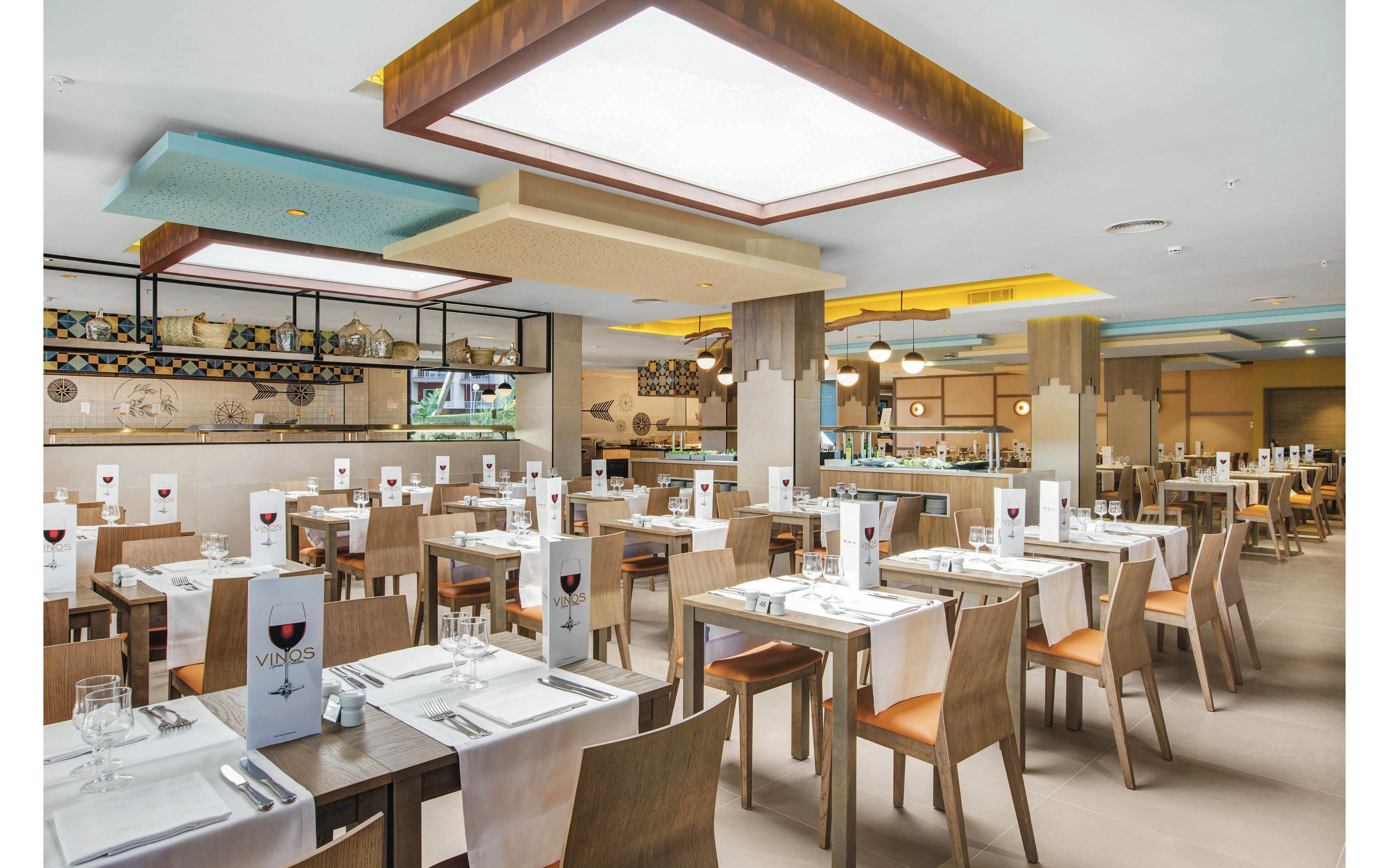 A spacious, contemporary restaurant dining room features neatly arranged wooden tables and chairs. Each table is set with white linens, glassware, and menus displaying the word 'VINOS' with a wine glass graphic. The setting is bright with large ceiling lights and a neutral color palette, creating a welcoming and clean atmosphere. The background shows a buffet area and decorative wall tiles.
