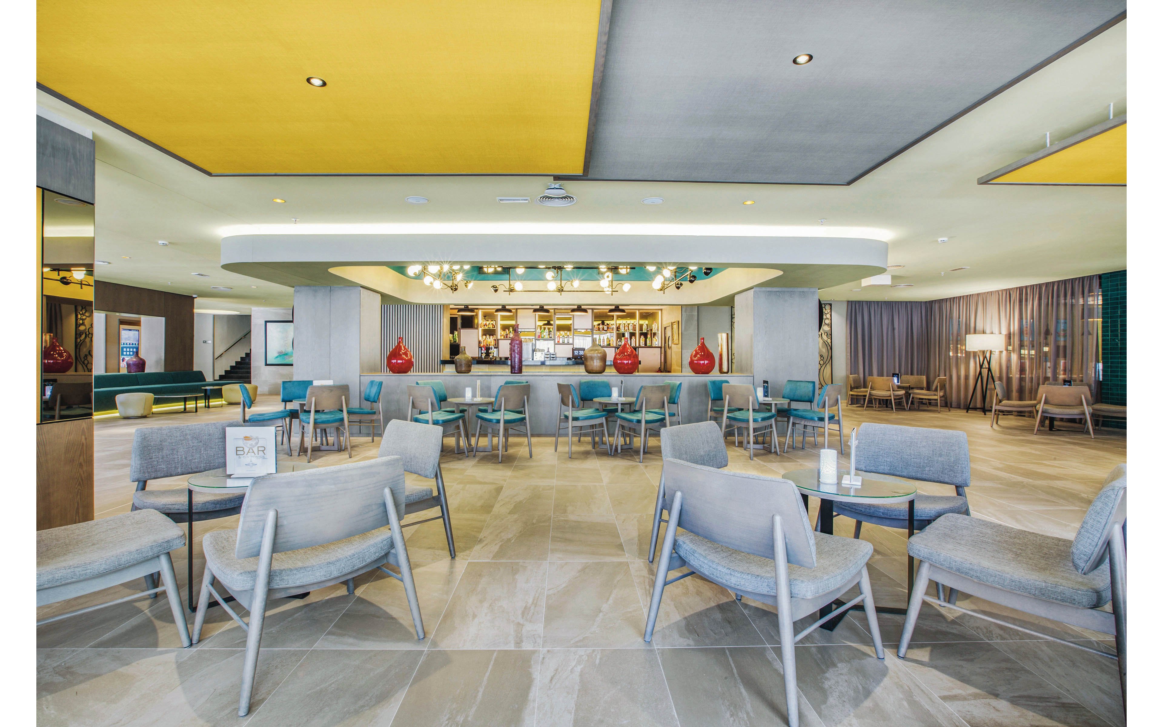 A spacious hotel lounge features a central bar with contemporary decor and ample seating. The setting includes light gray chairs, glass tables, and decorative red vases on the bar counter. The ceiling has bold yellow and gray panels, adding a vibrant touch to the neutral-toned flooring. The area is well-lit, with a relaxed and inviting atmosphere suitable for socializing or casual meetings.