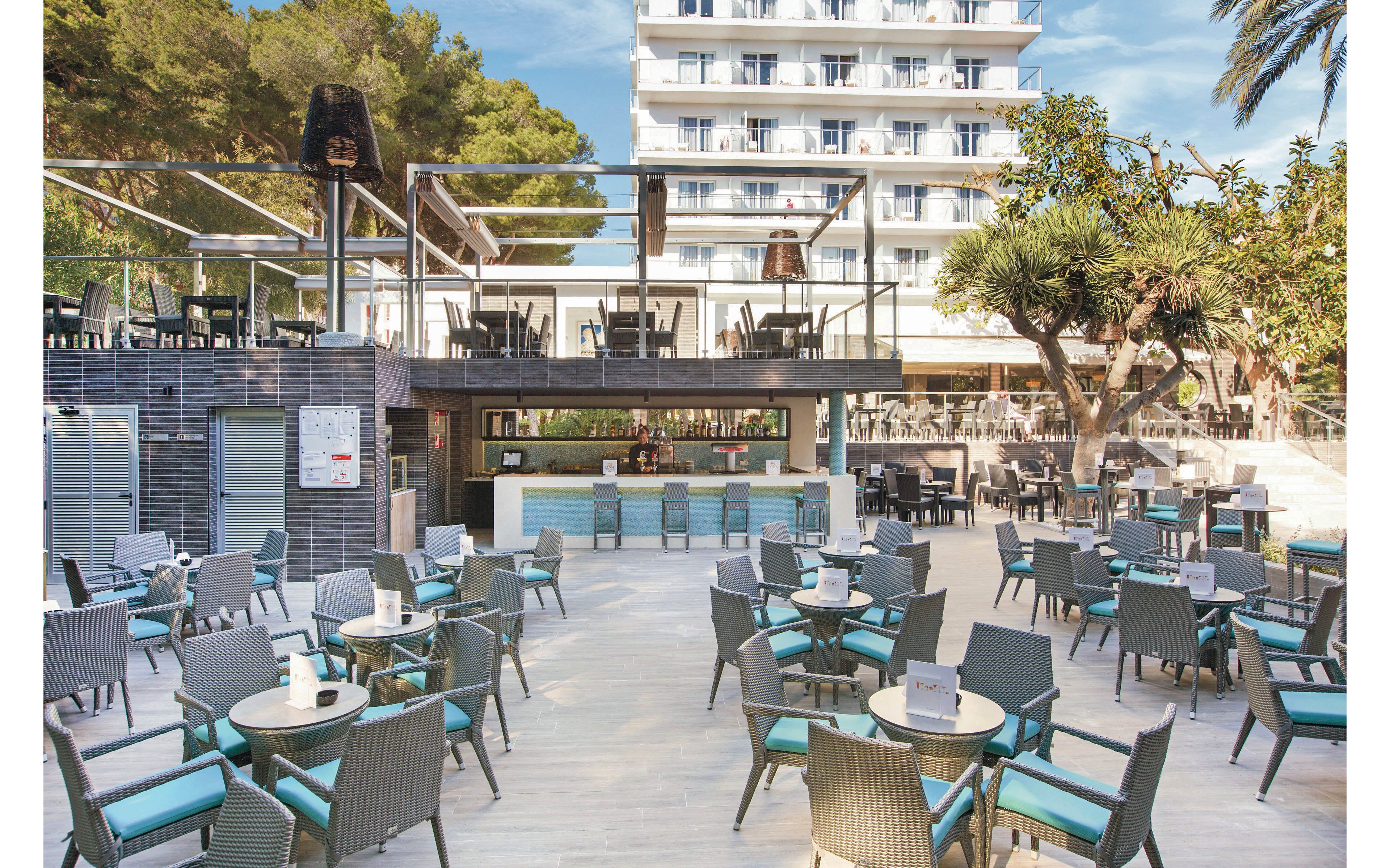 The image shows a spacious outdoor terrace at a hotel, featuring numerous tables and chairs arranged for guests. A central bar area is visible, with a modern design and clean lines. The setting is surrounded by trees and greenery, with a multi-story hotel building in the background. The overall mood is relaxed and inviting, with a palette of neutral tones and turquoise accents. No visible text or numeric values are present.