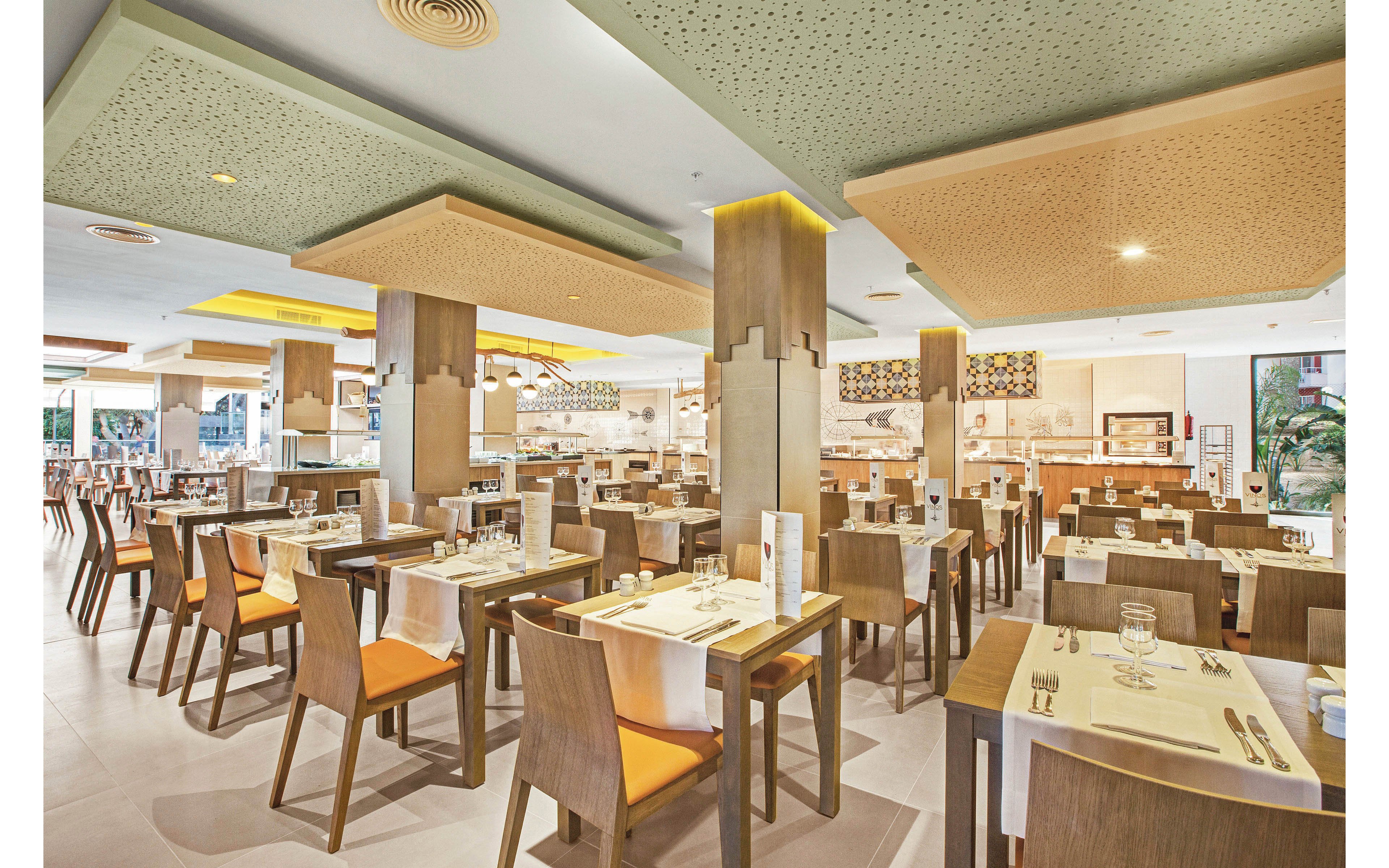 The image shows a spacious restaurant interior featuring neatly arranged wooden tables and chairs with orange cushions. The setting is bright and contemporary, with decorative ceiling panels and ample natural light. Table settings include glasses, cutlery, and menus, creating a welcoming atmosphere for guests. The overall mood is clean, inviting, and organized, suitable for casual or formal dining.