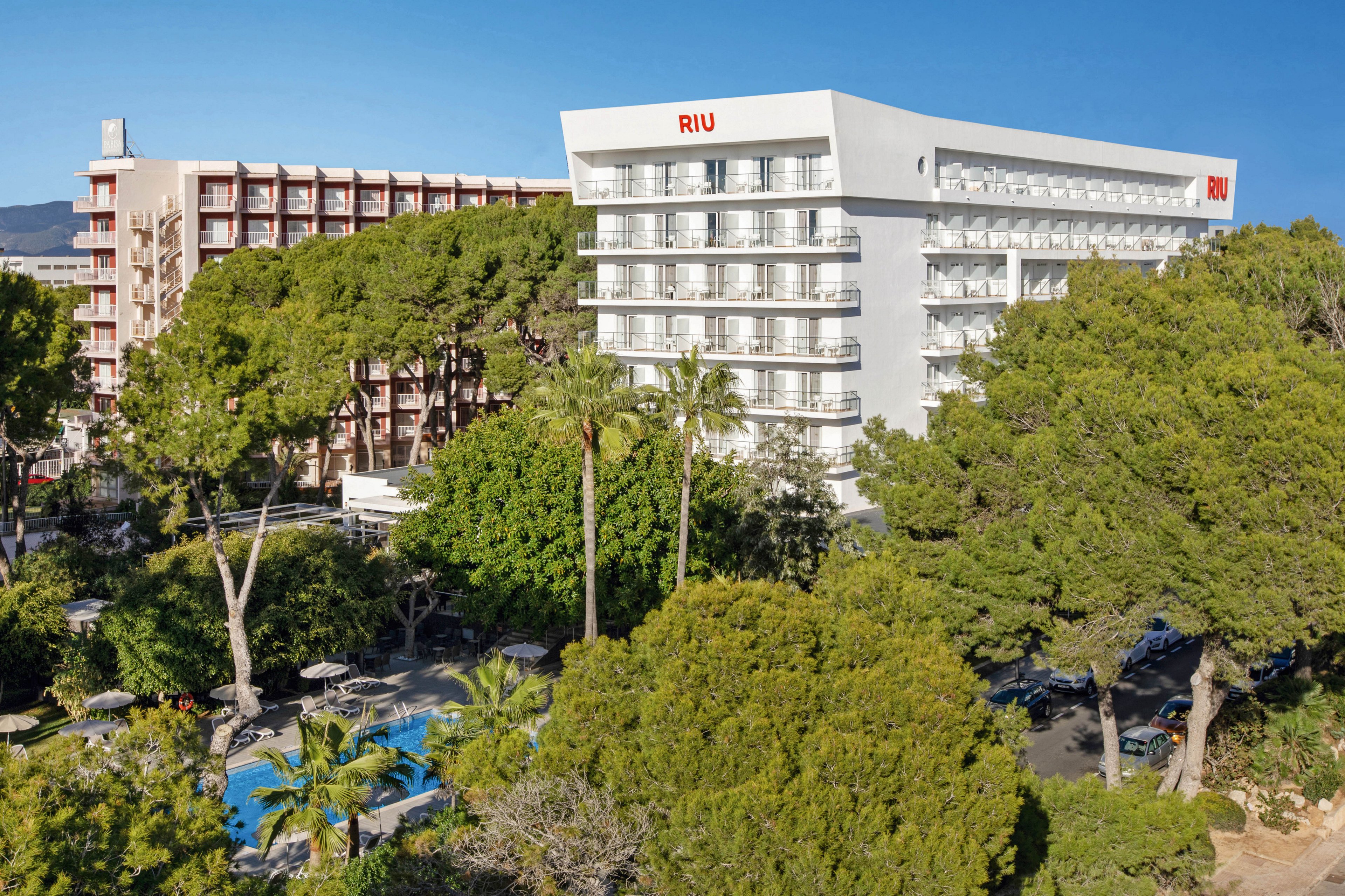 The image features the RIU hotel, clearly identified by the visible text on the building facade. The hotel is surrounded by tall trees and dense greenery, with a swimming pool area visible in the foreground. The setting appears to be a sunny day, creating a bright and inviting atmosphere. The architecture is modern, and the overall mood is relaxed and vacation-oriented.