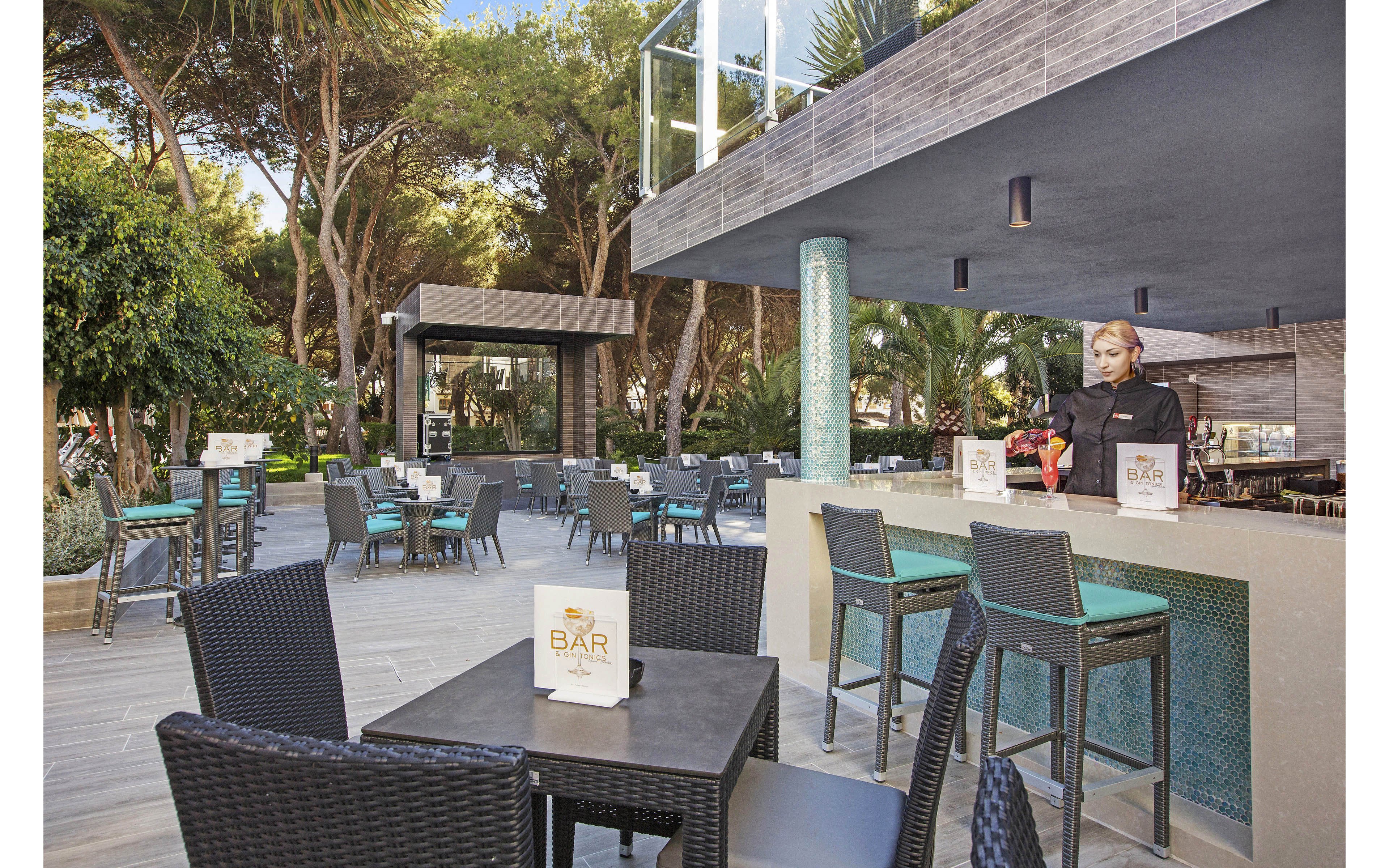 The image shows a contemporary outdoor bar setting with woven black chairs and tables, surrounded by lush greenery and tall trees. A bartender stands behind the bar counter, preparing drinks. Several tables display menus labeled 'BAR,' and the area is well-lit with natural daylight, creating a relaxed and inviting atmosphere. The space features turquoise accents and a clean, minimalist design.