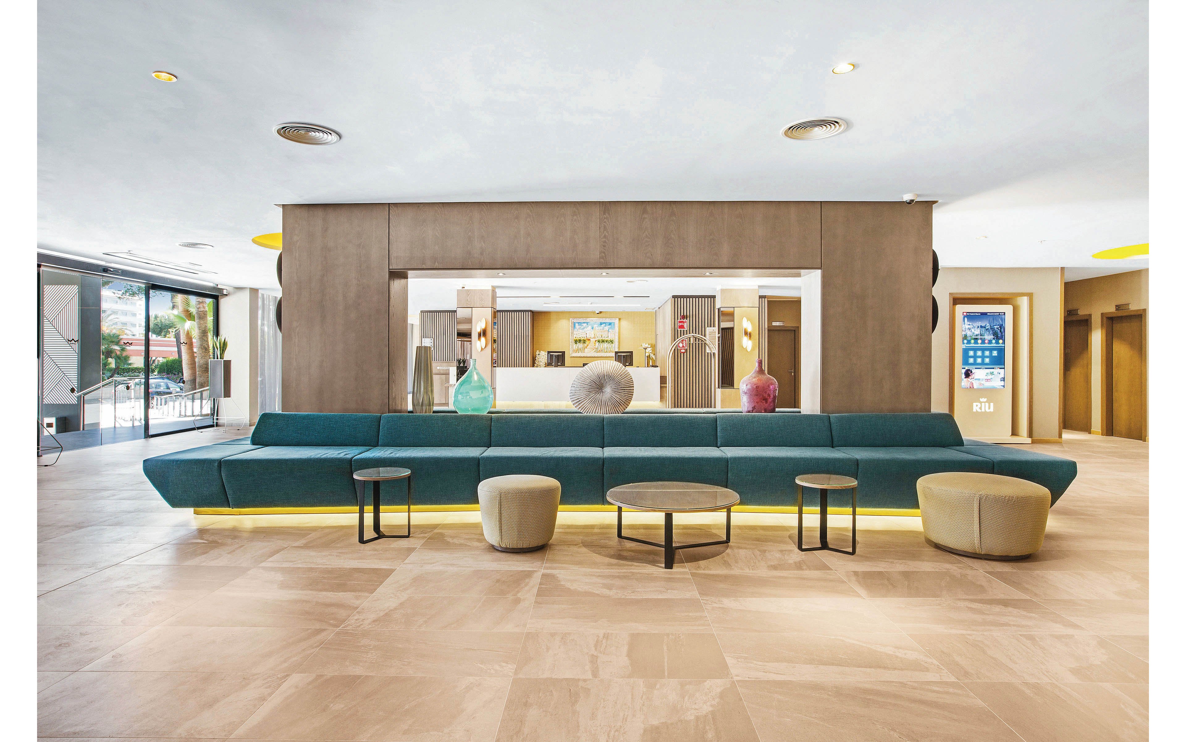 A spacious hotel lobby features a long teal sectional sofa with several round ottomans and tables in front. The setting is bright and contemporary, with neutral beige flooring and wood panel accents. Decorative vases and a reception desk are visible in the background, along with a digital display showing the number 24. The overall mood is welcoming and modern.