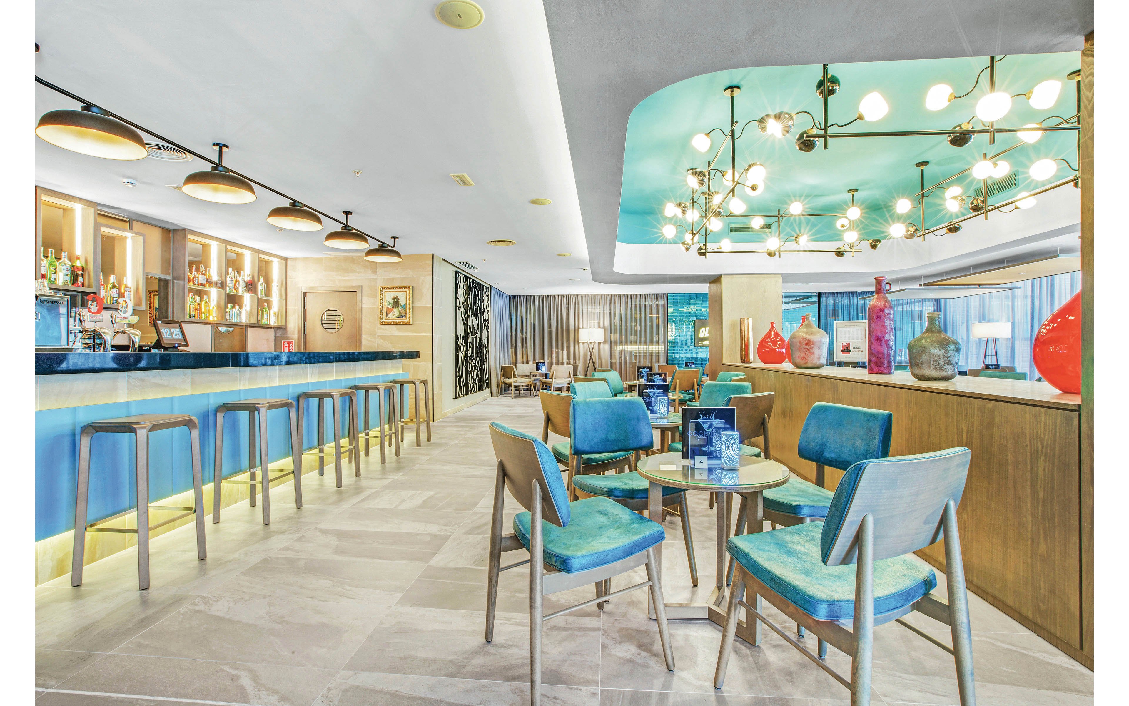 A contemporary hotel bar lounge features turquoise chairs and barstools, a sleek blue bar counter, and a ceiling with modern light fixtures. The setting is bright and inviting, with decorative vases and bottles displayed on wooden shelves and counters. The overall mood is stylish and relaxed, with a cool color palette and open layout.