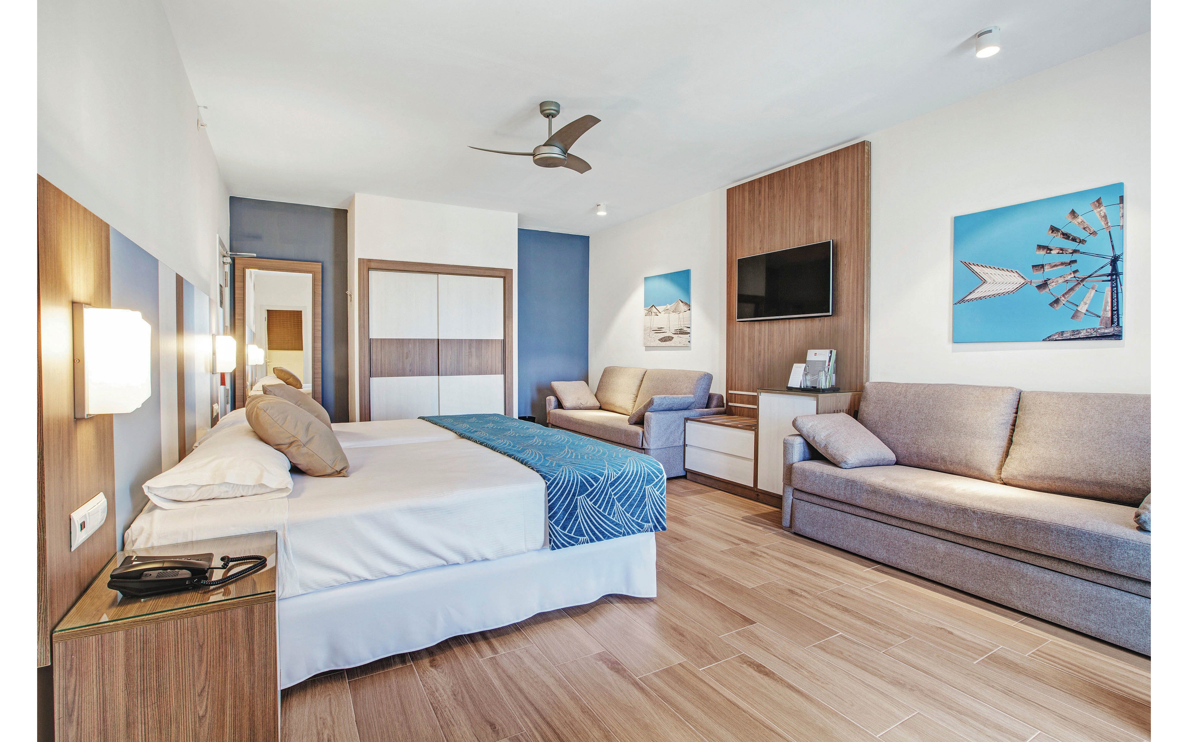 A spacious hotel room features a large double bed with white linens and a blue patterned runner. The room includes a sofa, armchair, wall-mounted television, and contemporary wooden furnishings. Light wood flooring, neutral tones, and blue accent walls create a calm, modern atmosphere. Framed artwork and a ceiling fan add to the inviting setting.