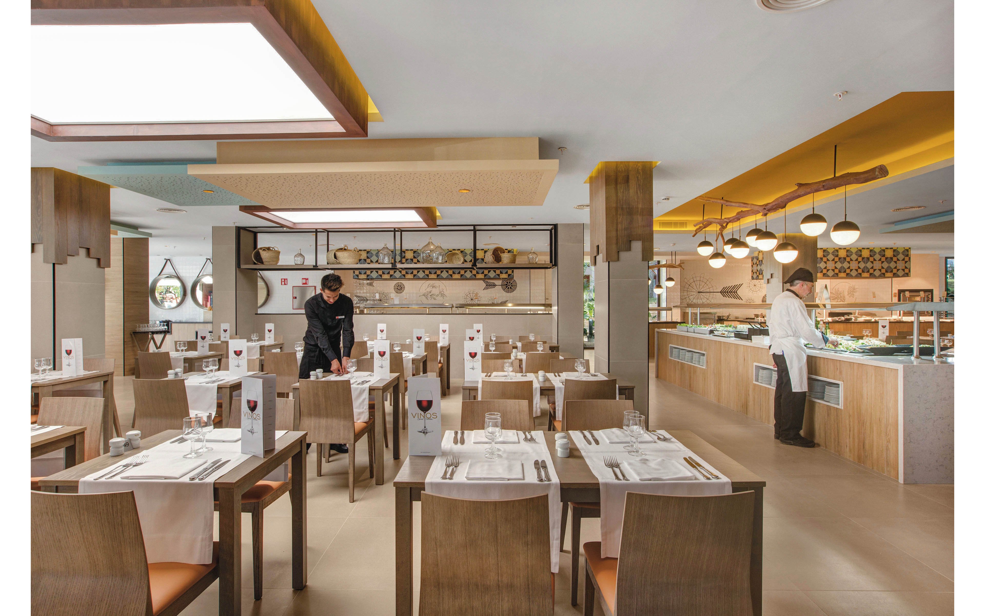 A contemporary restaurant interior features neatly arranged tables and chairs, with two staff members preparing the dining area. The setting is bright and spacious, with warm lighting and a buffet counter visible in the background. Table settings include menus and glassware, and the overall palette is neutral with wood accents. The atmosphere appears calm and organized, suitable for casual or family dining.