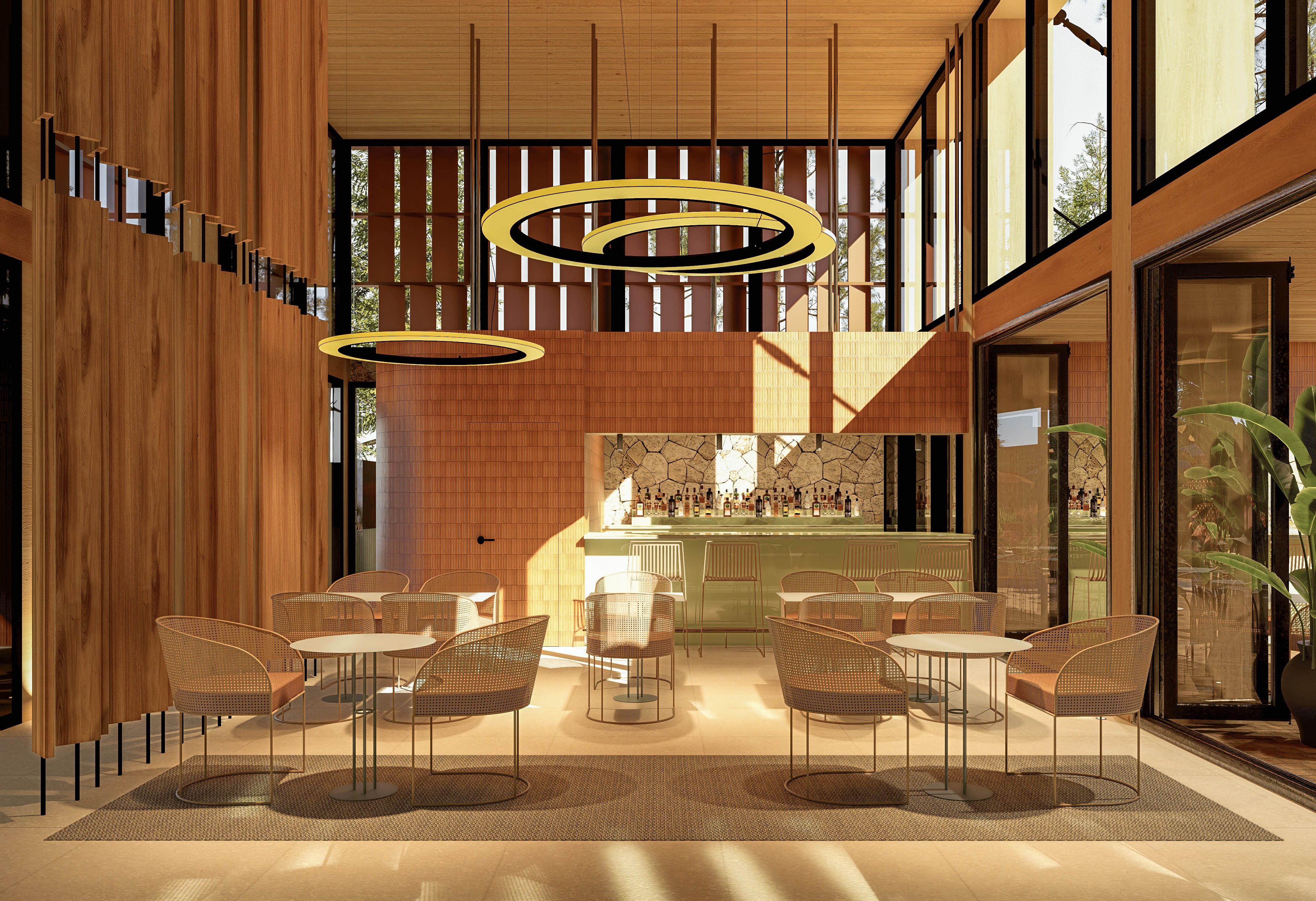 A contemporary café interior features a central bar area with light green cabinetry and patterned tile backsplash. Circular ring pendant lights hang from a high wooden ceiling, casting warm illumination over the space. Woven chairs surround round tables on a large rug, creating an inviting seating area. Floor-to-ceiling windows and wood panel walls allow natural light to flood the room and reveal greenery outside.