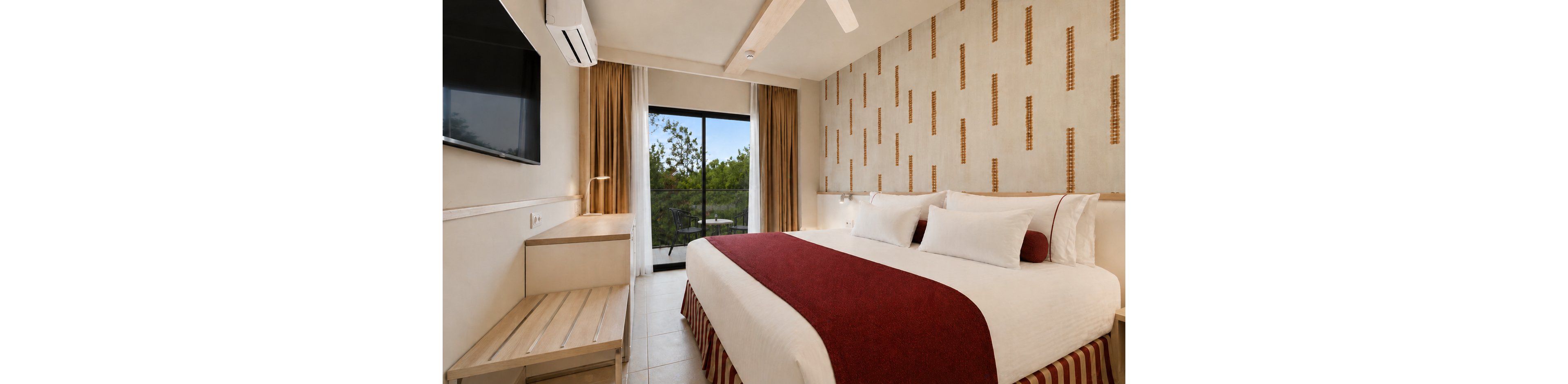 A contemporary hotel bedroom features a large bed with white linens and a red runner. The room includes a wall-mounted flat-screen television, a light wooden desk, and a bench. Floor-to-ceiling curtains frame a sliding glass door that opens to a balcony with greenery outside. Neutral walls with a subtle patterned accent and a ceiling fan contribute to a calm, minimalist atmosphere.