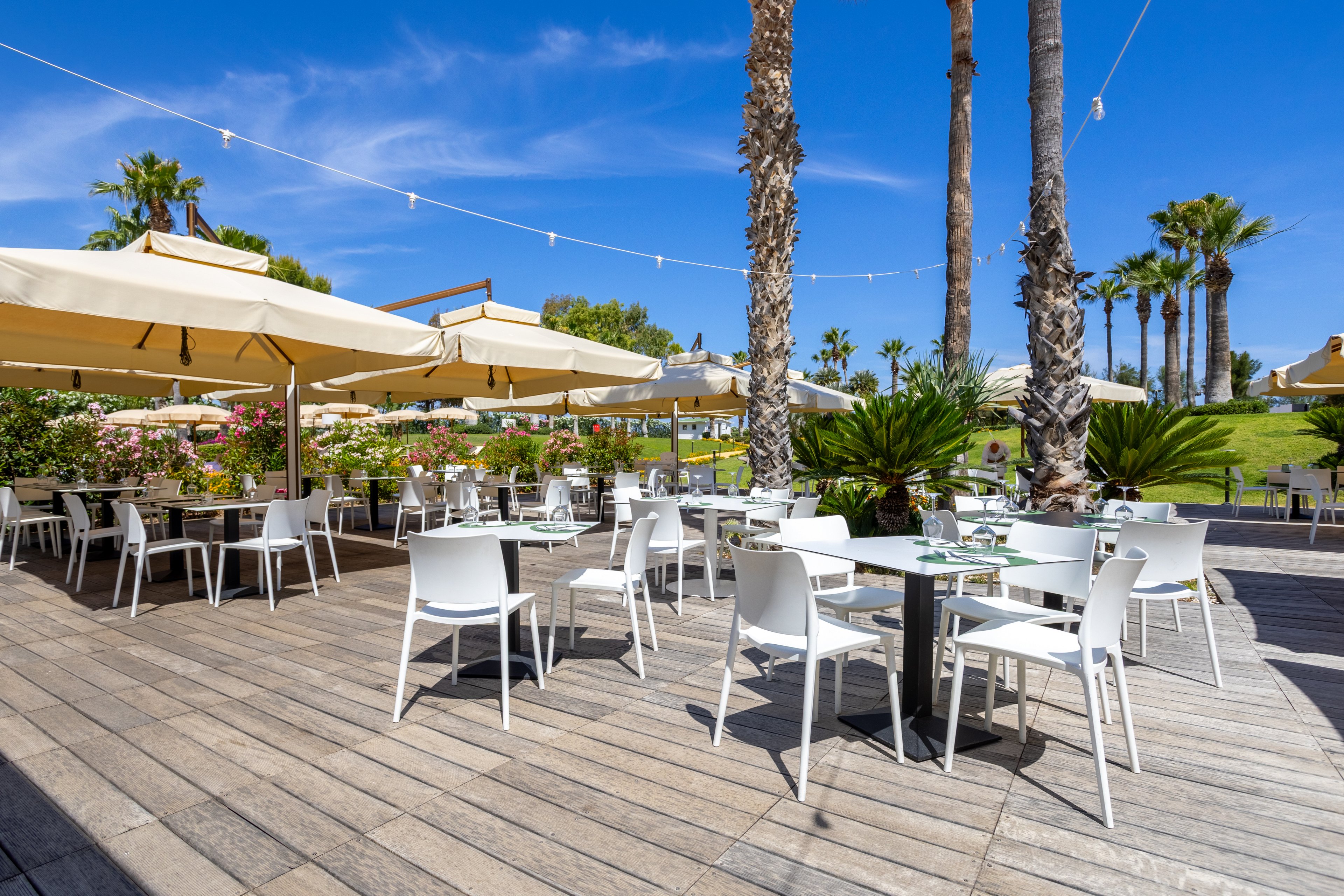 An open-air restaurant terrace features white tables and chairs neatly arranged under large beige sun umbrellas. The setting is bright and sunny, with palm trees, manicured lawns, and flowering shrubs surrounding the decked area. Glassware and table settings are prepared, suggesting readiness for guests in a relaxed vacation atmosphere. A clear blue sky with string lights overhead adds to the inviting, resort-like ambiance.