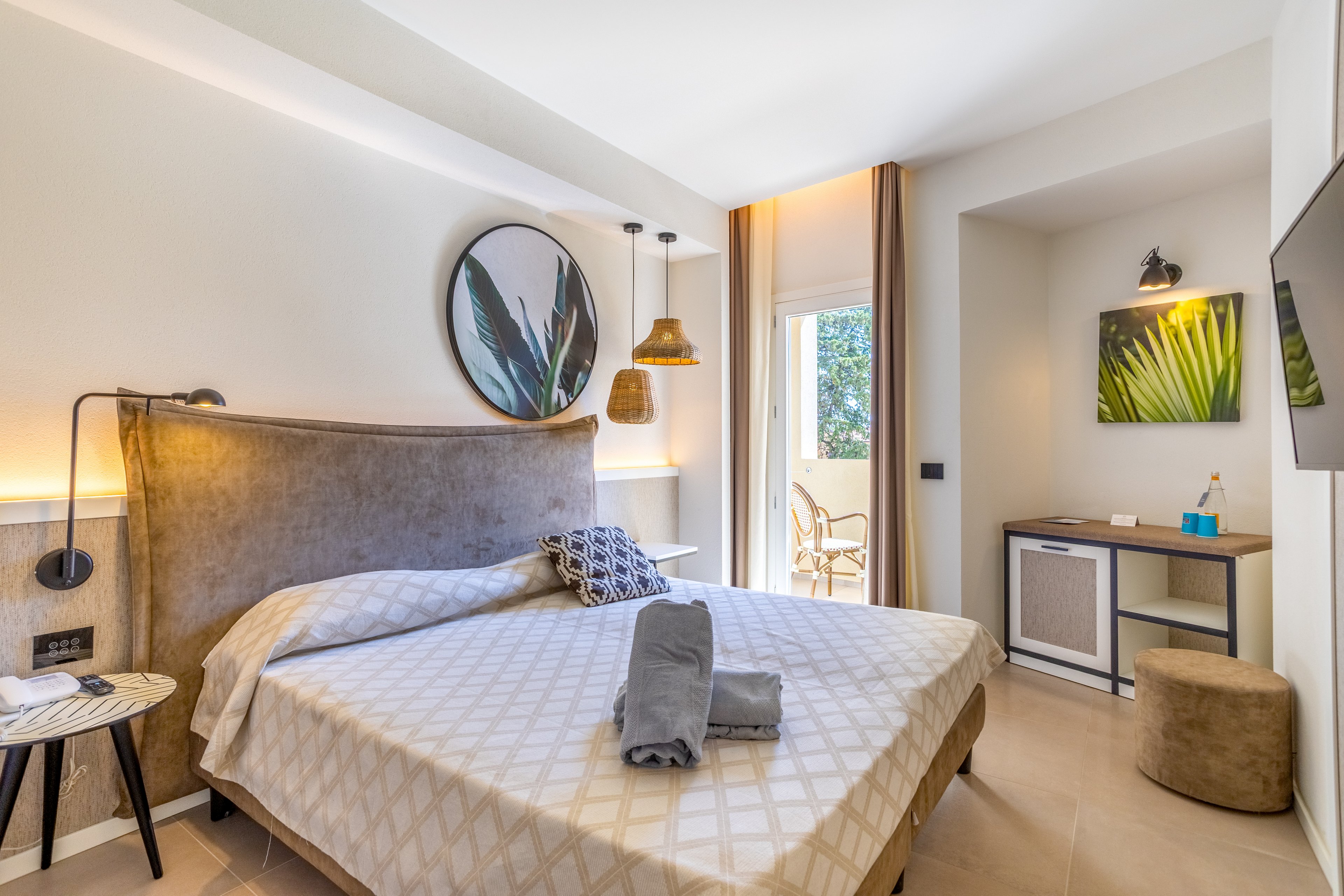 Contemporary hotel bedroom featuring a large bed with patterned bedding and neatly folded towels at the foot. The room is decorated in warm neutral tones with botanical wall art and woven pendant lights. A small desk area with a chair and stool sits opposite the bed, next to a wall-mounted television. Natural light streams in from the open balcony door, creating an inviting and relaxing atmosphere.