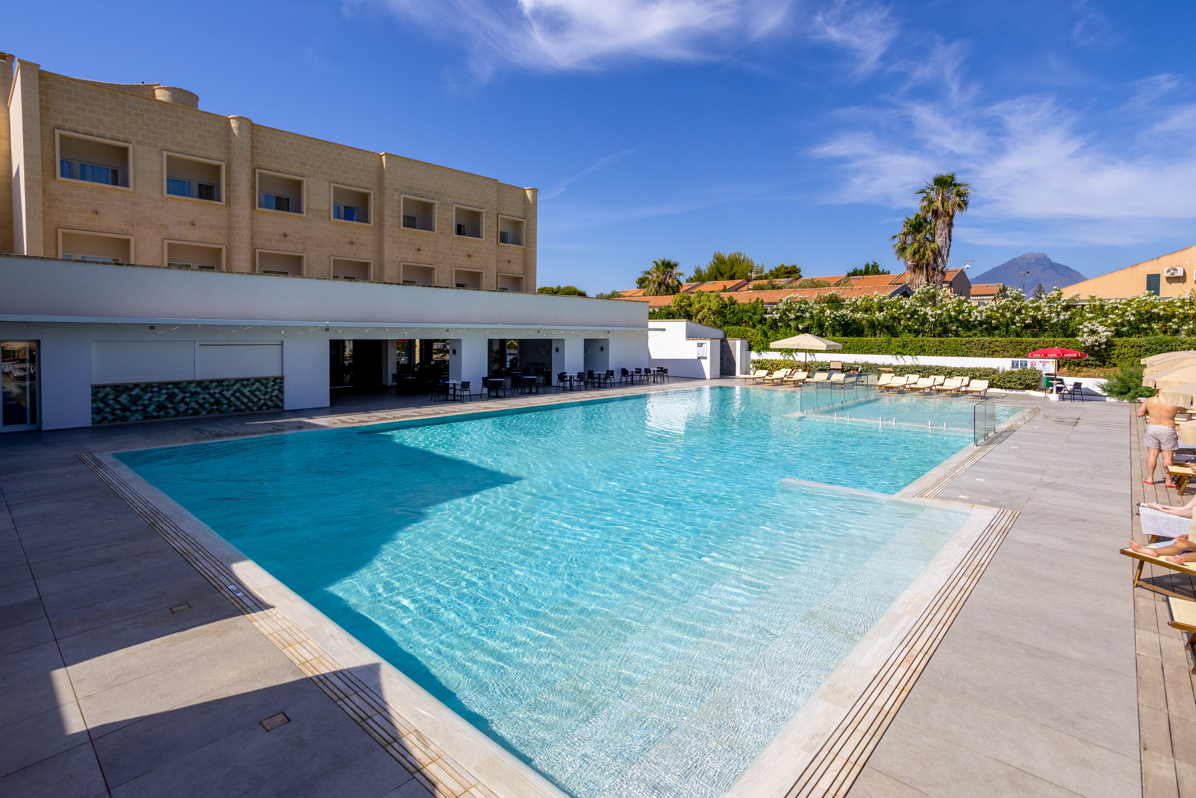 Large turquoise swimming pool sits in front of a modern hotel building under a clear blue sky. Sun loungers and umbrellas line the far side of the pool, suggesting a relaxing vacation setting. Palm trees and distant mountains frame the background, adding a Mediterranean or resort feel. Bright daylight and clean architectural lines create a fresh, inviting atmosphere.