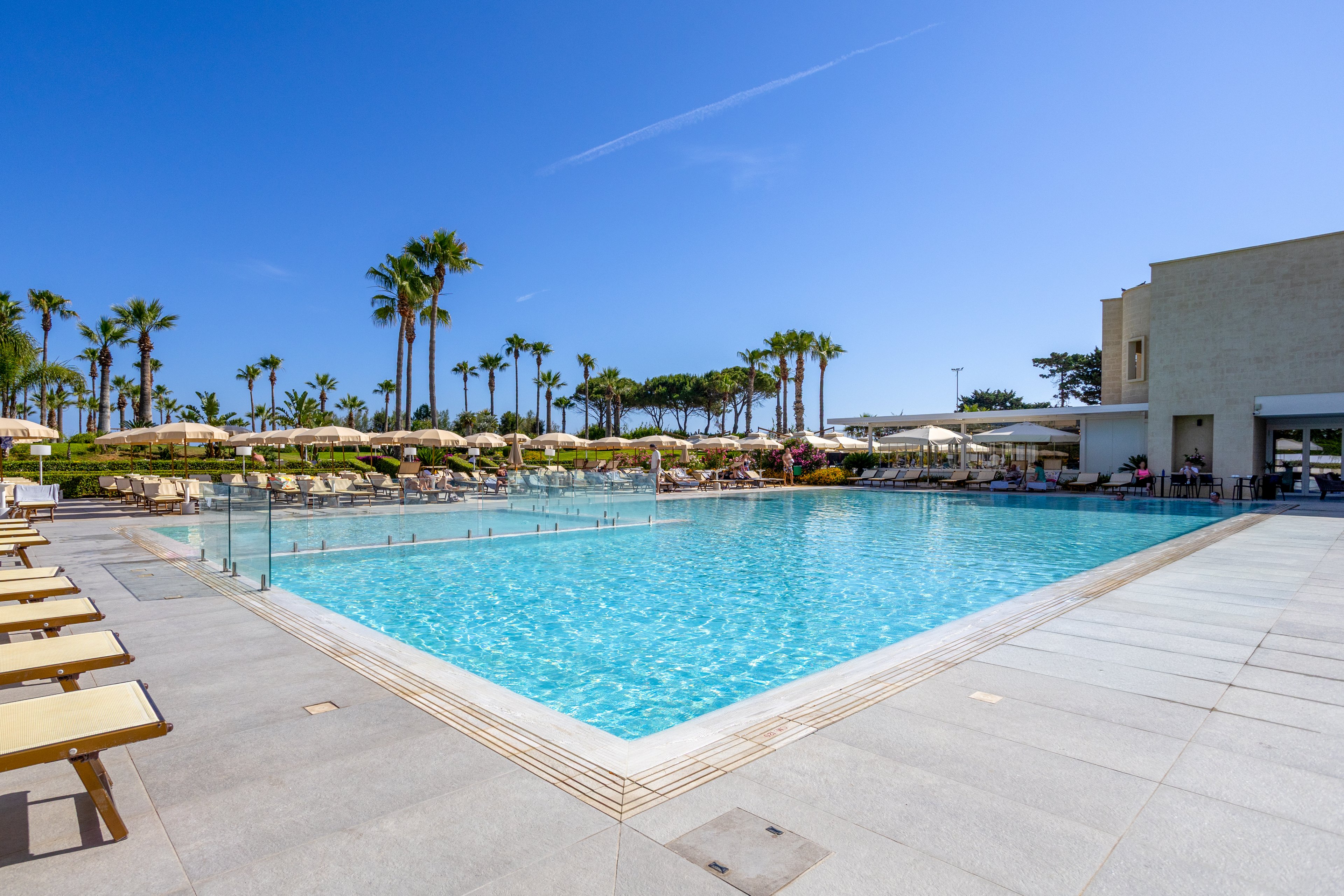 A large outdoor swimming pool is surrounded by sun loungers and umbrellas at a modern resort. Palm trees line the background, adding a tropical feel to the bright, sunny setting. The clear blue water contrasts with the light stone deck and contemporary buildings. The scene suggests relaxation, vacation leisure, and warm weather.