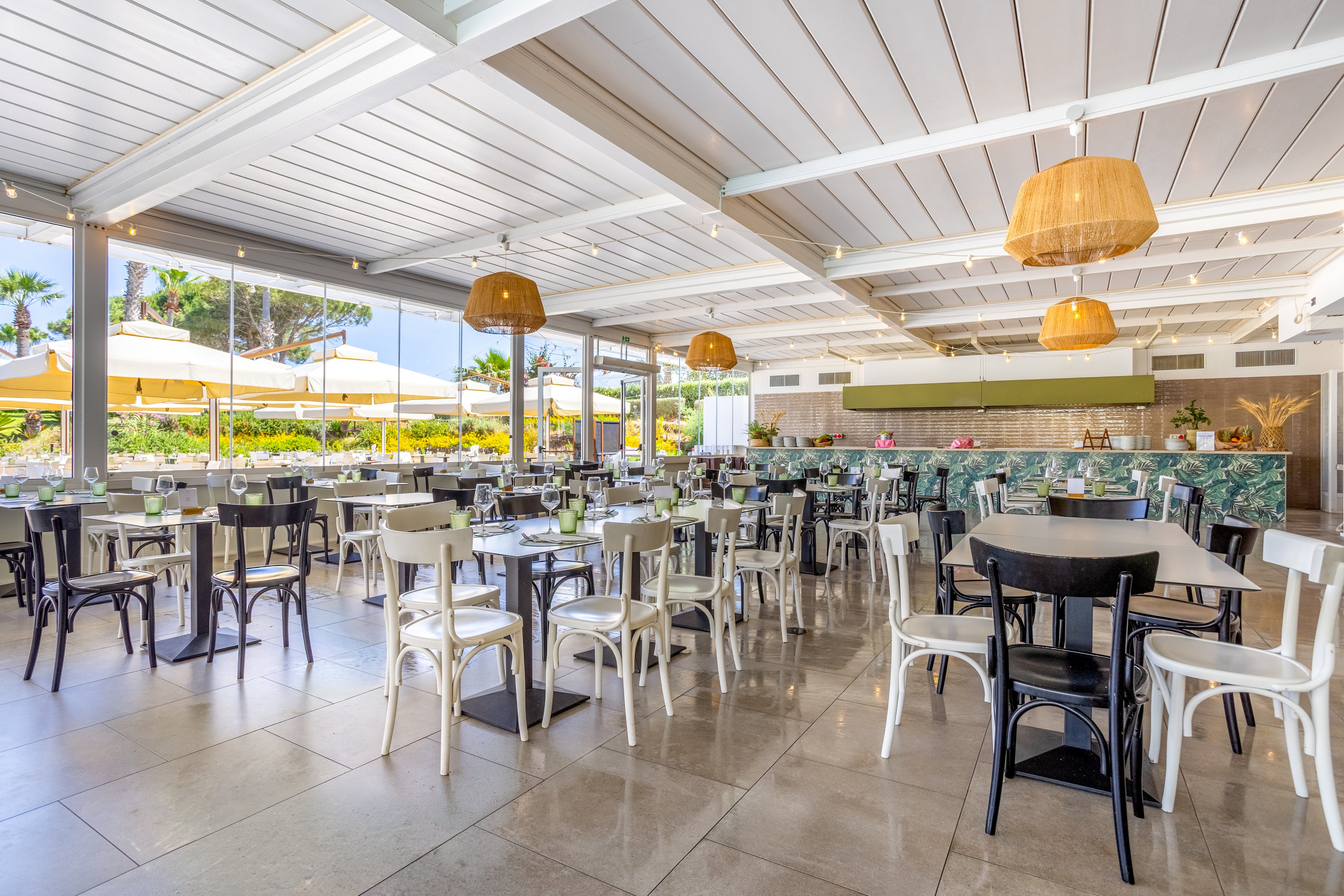 Spacious open-air restaurant interior with neatly arranged tables and chairs. Large windows reveal a sunny outdoor terrace with yellow umbrellas and lush greenery. Warm wicker pendant lights hang from the white ceiling, adding a relaxed, modern touch. The setting feels inviting and casual, ideal for daytime dining in a coastal or resort environment.