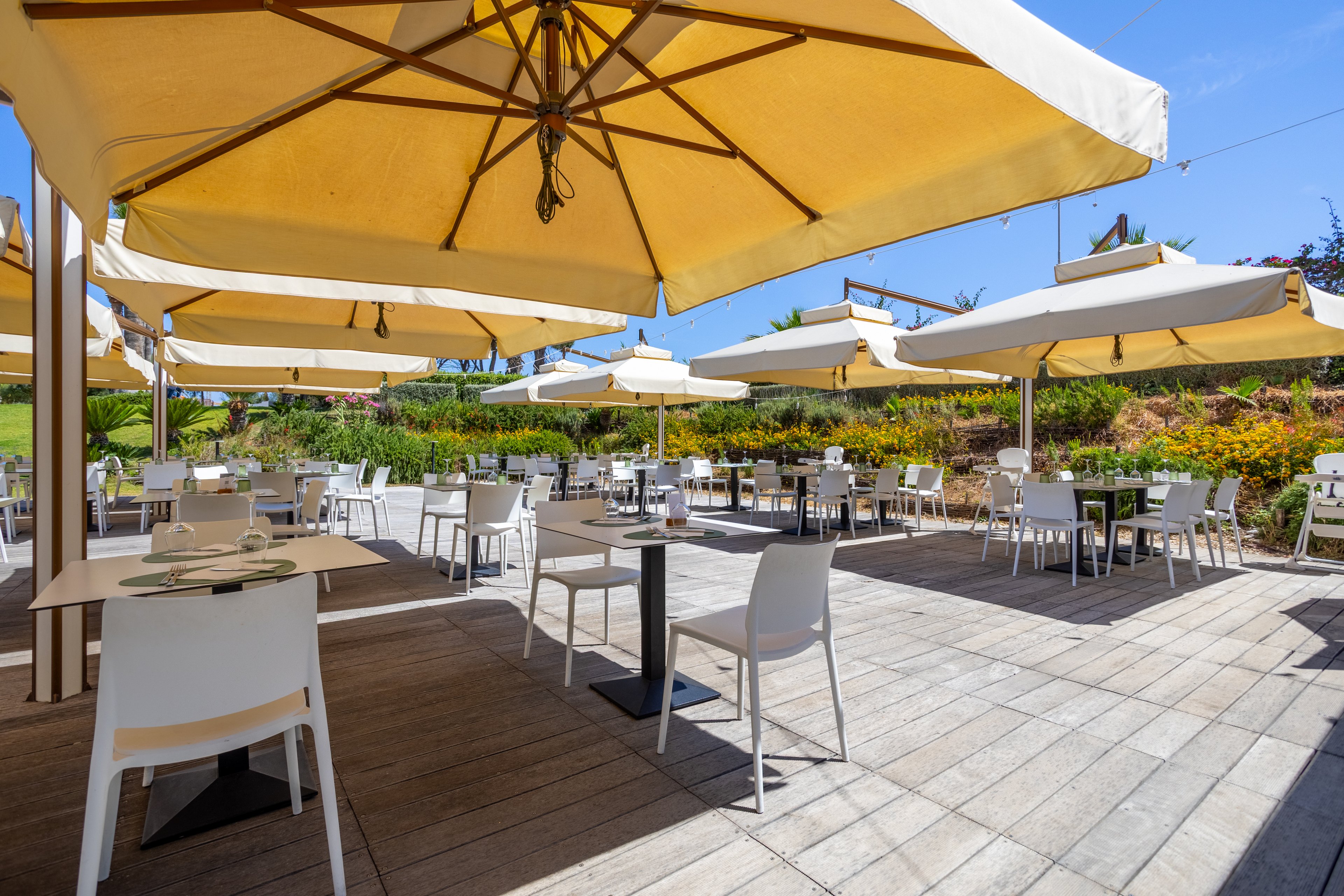 An open-air restaurant terrace features neatly arranged white tables and chairs under large beige sun umbrellas. The setting appears bright and sunny, with clear blue sky and lush greenery in the background. Place settings are prepared on several tables, suggesting readiness for guests in a relaxed, casual dining atmosphere. The spacious layout and wooden deck flooring create an inviting environment for outdoor meals.