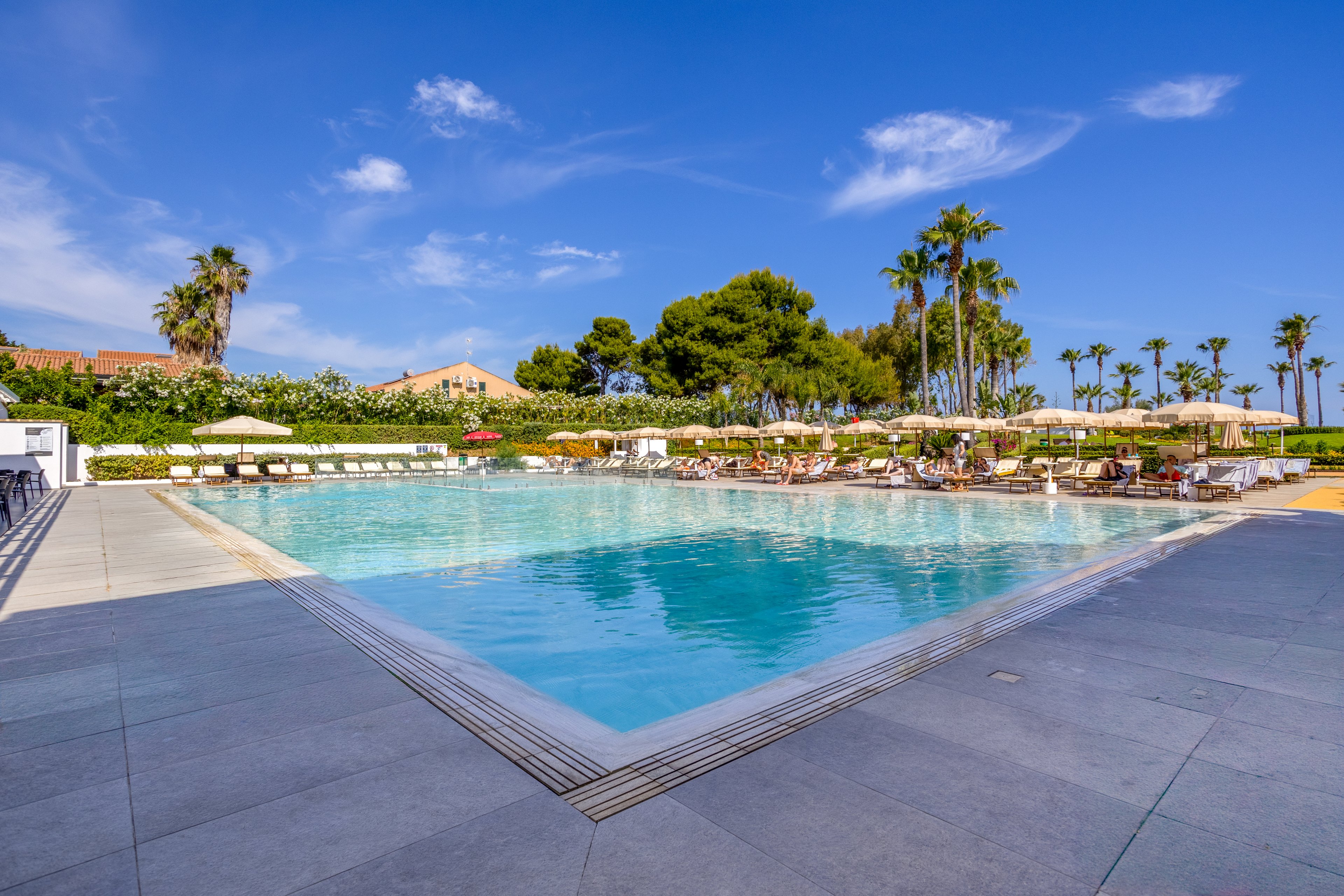 A large outdoor swimming pool is surrounded by sun loungers and umbrellas at a resort. People relax by the water under a bright blue sky with scattered clouds. Palm trees and lush greenery frame the pool area, creating a tropical vacation atmosphere. The scene feels calm and inviting, ideal for leisure and summer holidays.