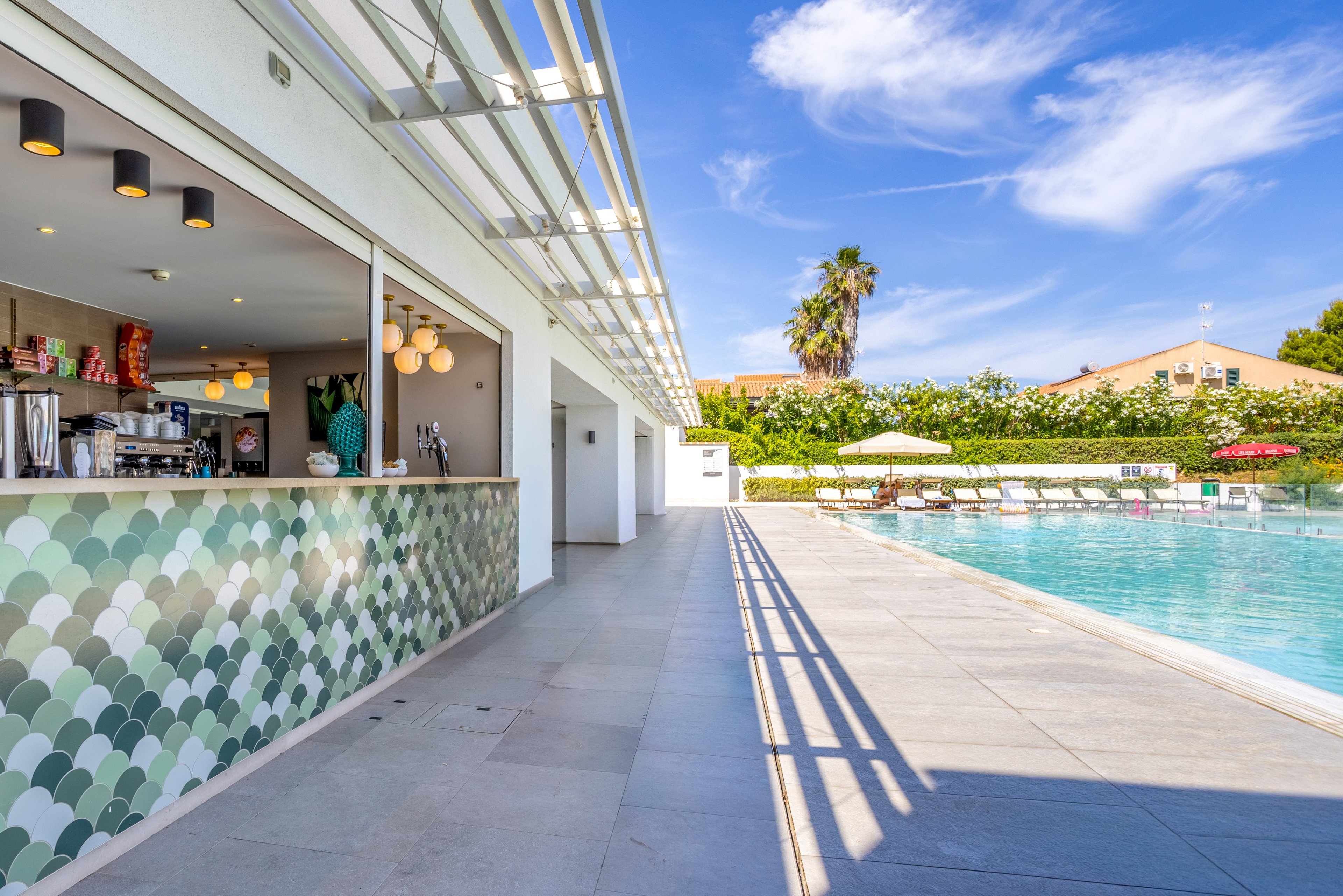 An open-air bar with a patterned tile counter sits beside a large outdoor swimming pool. Sun loungers and umbrellas line the far side of the pool, framed by lush greenery and low residential-style buildings. Bright daylight and a clear blue sky create a relaxed vacation atmosphere. The scene suggests leisure, refreshments, and poolside relaxation at a modern resort.