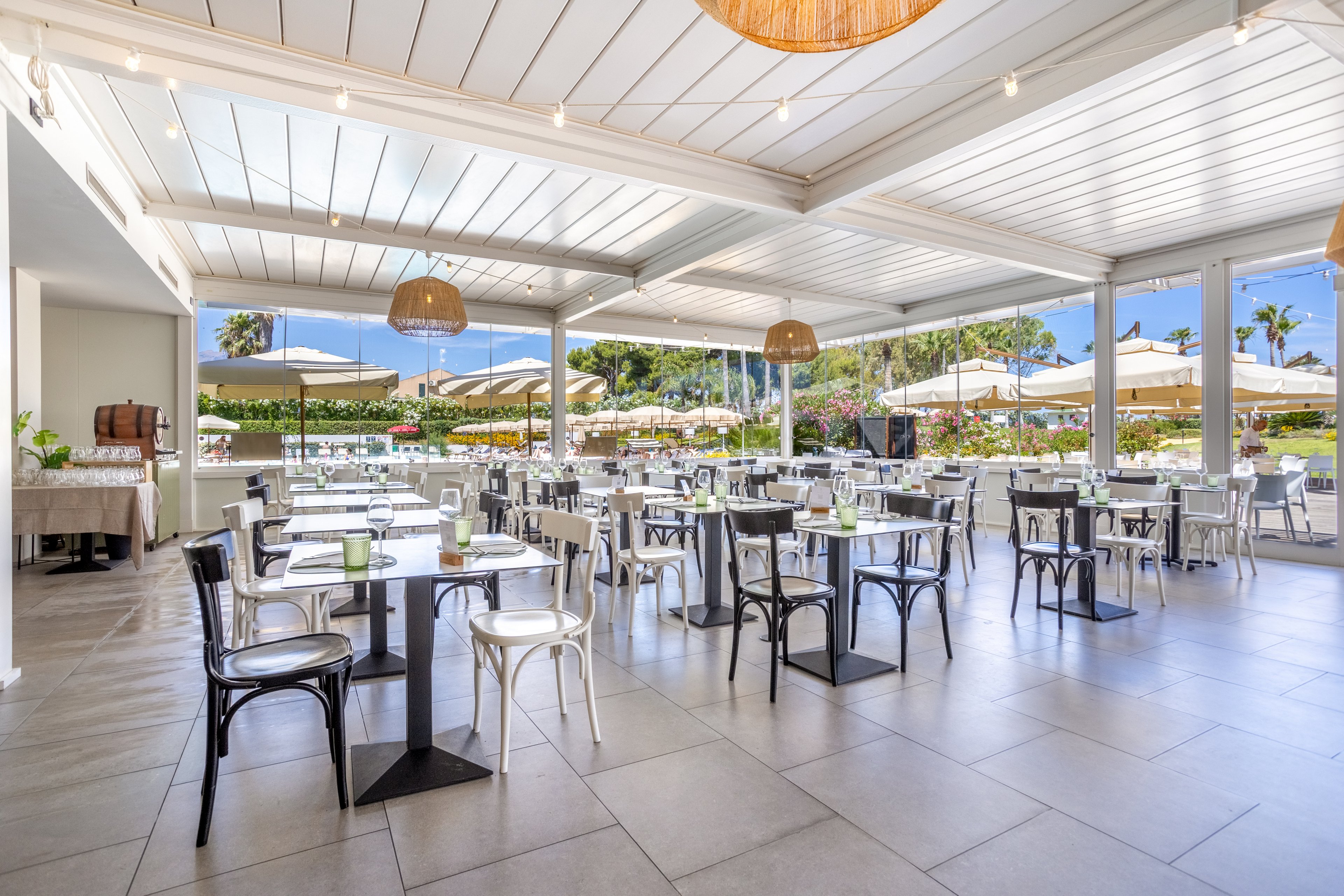 Bright open-air restaurant dining area with neatly arranged tables and chairs. The space features a modern covered terrace overlooking a lush garden and poolside umbrellas. Natural light floods the interior, highlighting neutral tones and simple contemporary decor. The setting appears calm and inviting, suitable for vacation dining or events.