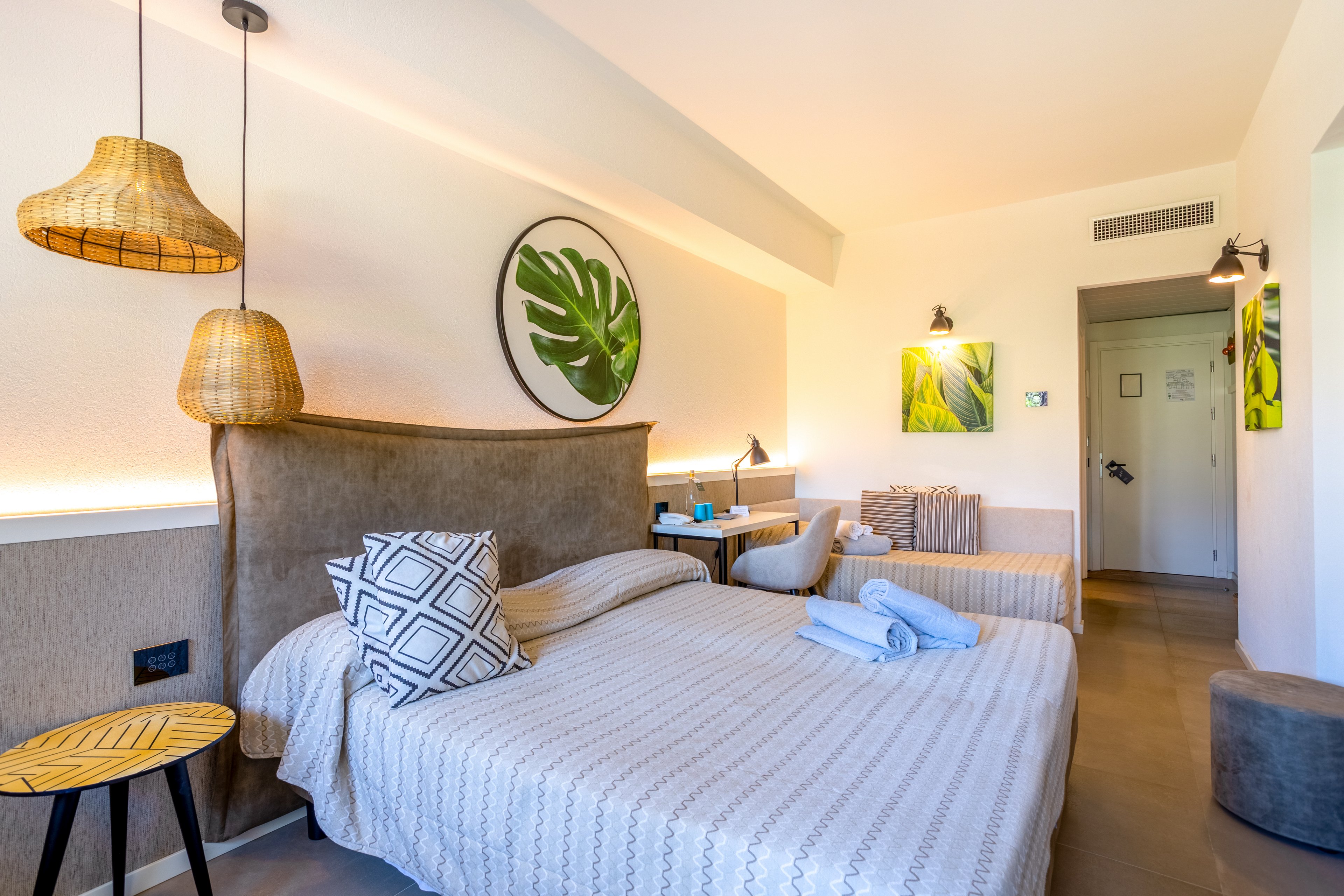A contemporary hotel room features two neatly made twin beds with neutral striped bedding and decorative pillows. The space is warmly lit by woven pendant lamps and wall sconces, creating a cozy and inviting atmosphere. Tropical leaf artwork adorns the walls, adding a fresh, natural accent to the minimalist decor. Rolled towels are placed on the bed, and a small seating area and desk are visible in the background.