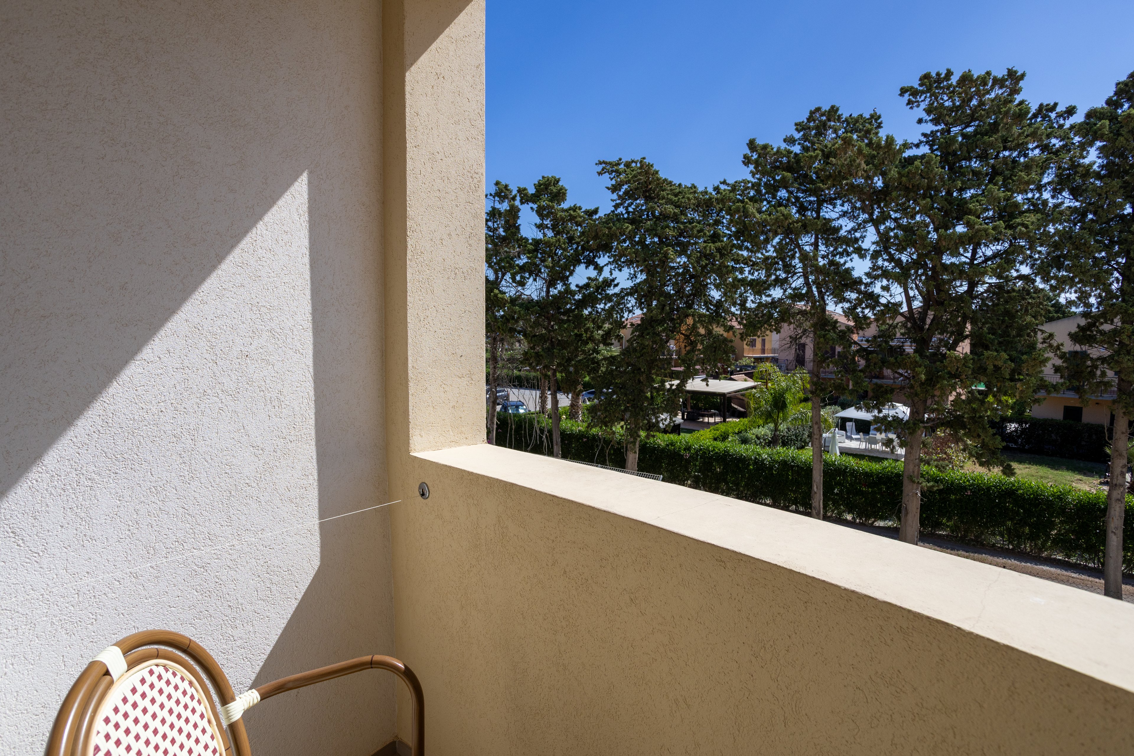 A small balcony with a light-colored wall and a single chair is shown in bright sunlight. Beyond the balcony, tall green trees line a quiet residential street. Houses and garden areas are partially visible through the foliage, suggesting a peaceful neighborhood setting. The clear blue sky enhances the warm, relaxed atmosphere of the outdoor space.