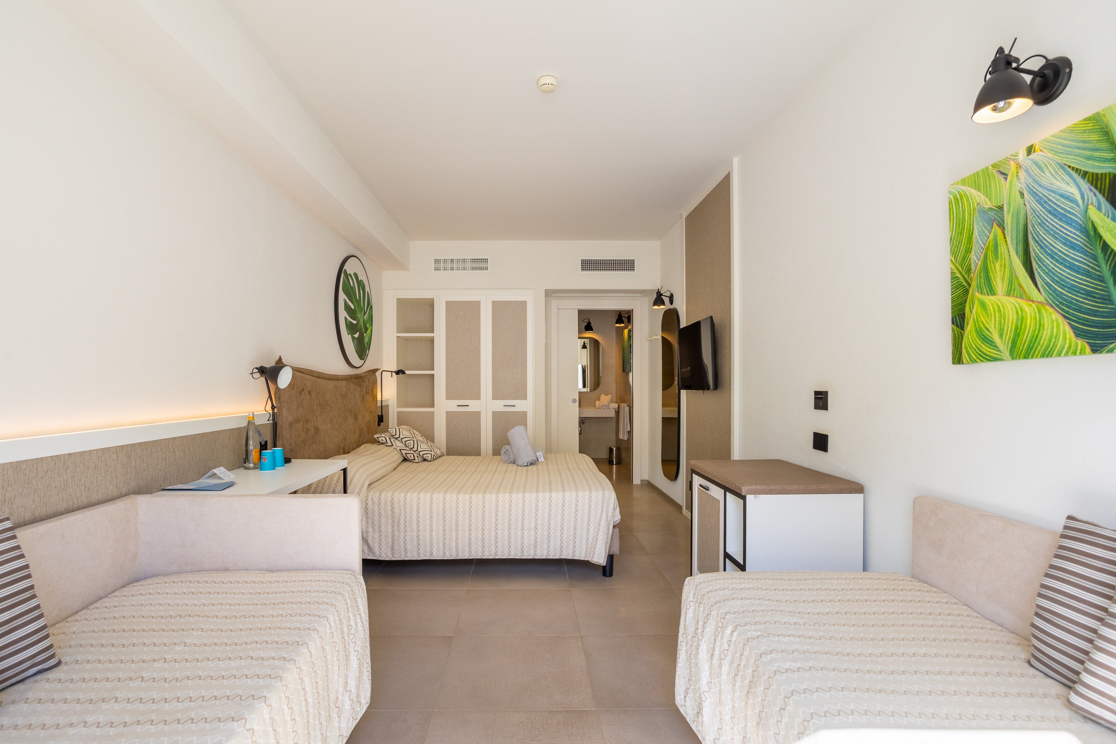 A bright, modern hotel room features a double bed in the center and two single beds on either side. Neutral tones dominate the decor, complemented by striped cushions and light-colored flooring. A wall-mounted television, small desk, and bedside lamps add functional comfort to the space. Green leaf artwork and natural light from an unseen window create a fresh, inviting atmosphere.