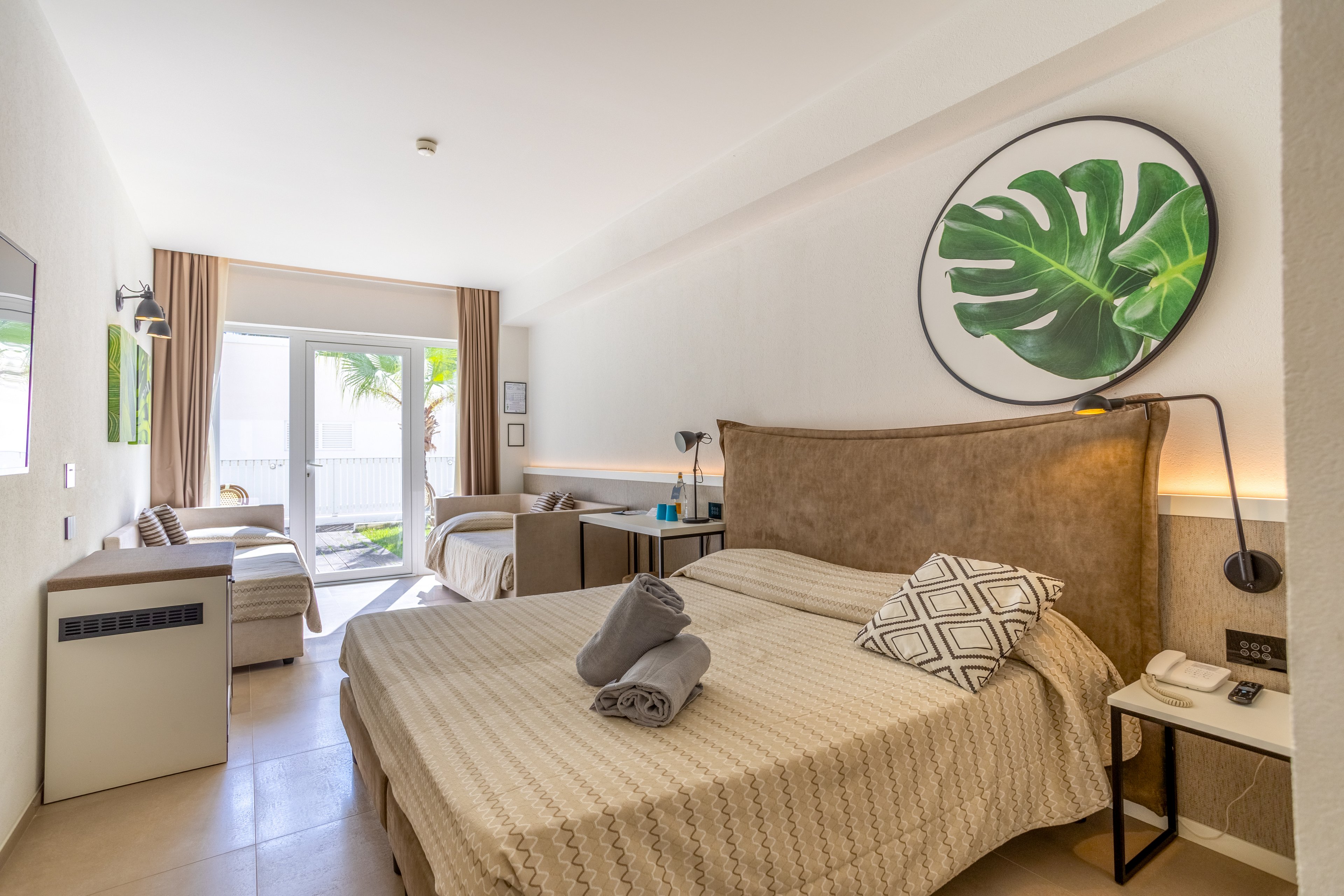 A contemporary hotel room features a large bed with folded towels and patterned cushions in the foreground. Neutral tones and minimalist furniture create a calm, inviting atmosphere. A circular wall art piece with a green leaf adds a fresh accent above the headboard. Sunlight streams through glass doors that open to an outdoor terrace with palm trees visible outside.