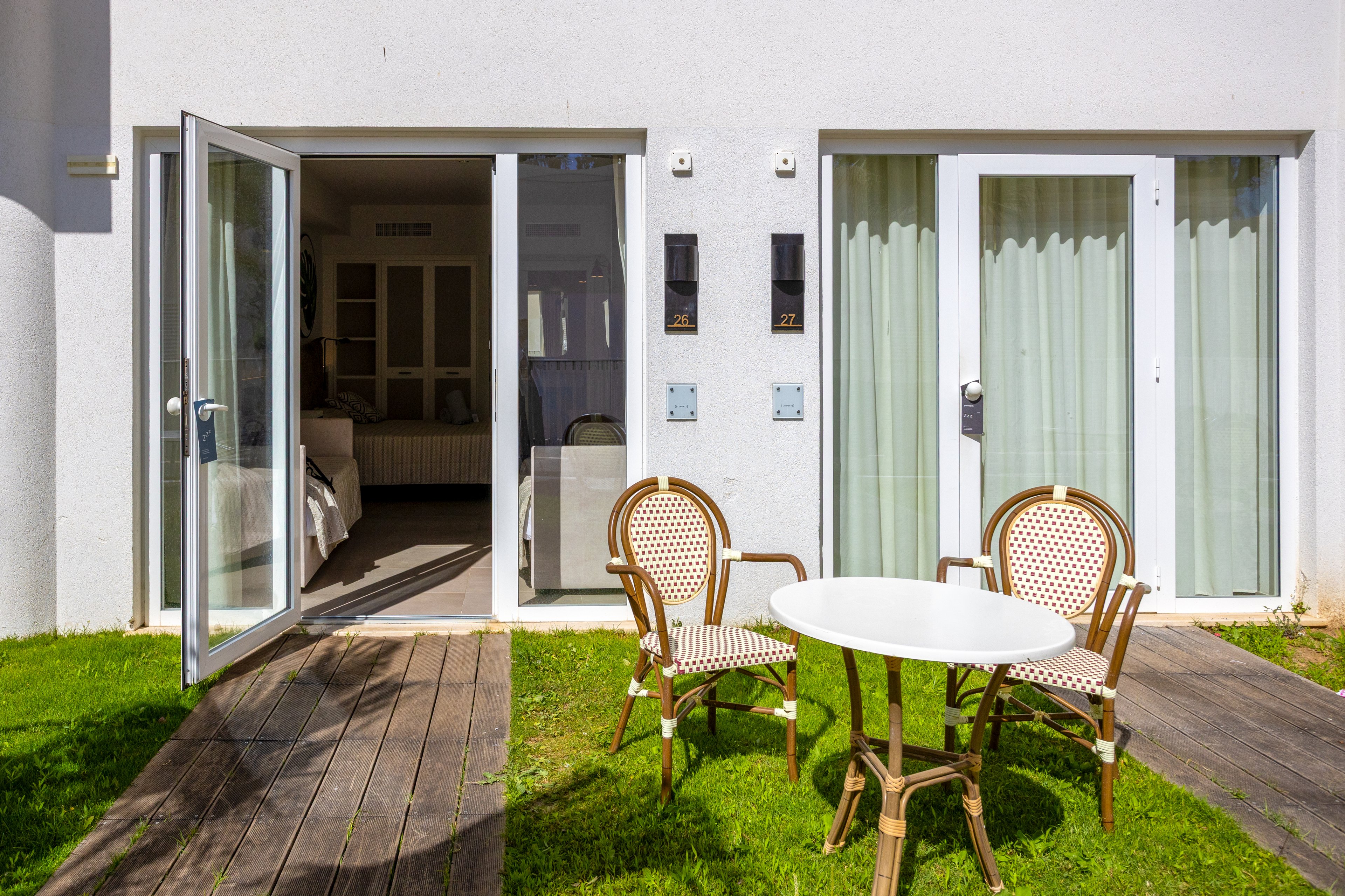 A small outdoor patio with two patterned chairs and a round white table sits on a grassy area beside a modern building. Large glass doors open into a bright bedroom with a neatly made bed visible inside. Wooden decking leads from the grass to the doorway, and sheer curtains cover the adjacent closed door. The scene is lit by strong sunlight, creating a warm and inviting atmosphere.