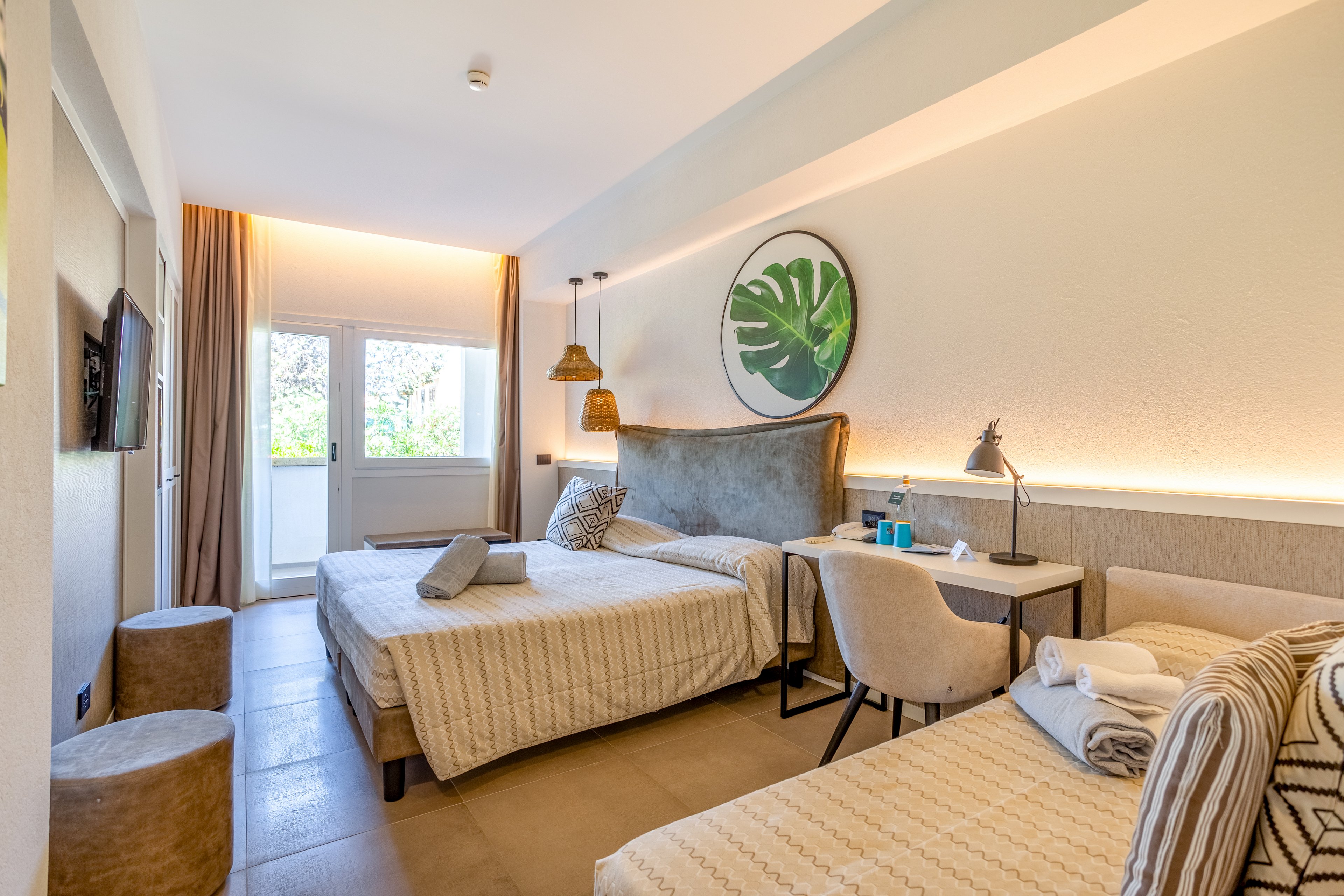 A contemporary hotel room features two neatly made beds with neutral bedding and accent pillows. The space is bright, with natural light streaming in from a balcony door and window at the far end. Warm ambient lighting, woven pendant lamps, and a large circular wall art with a green leaf create a cozy atmosphere. A desk with chairs, folded towels, and small amenities add to the welcoming, vacation-ready feel of the room.
