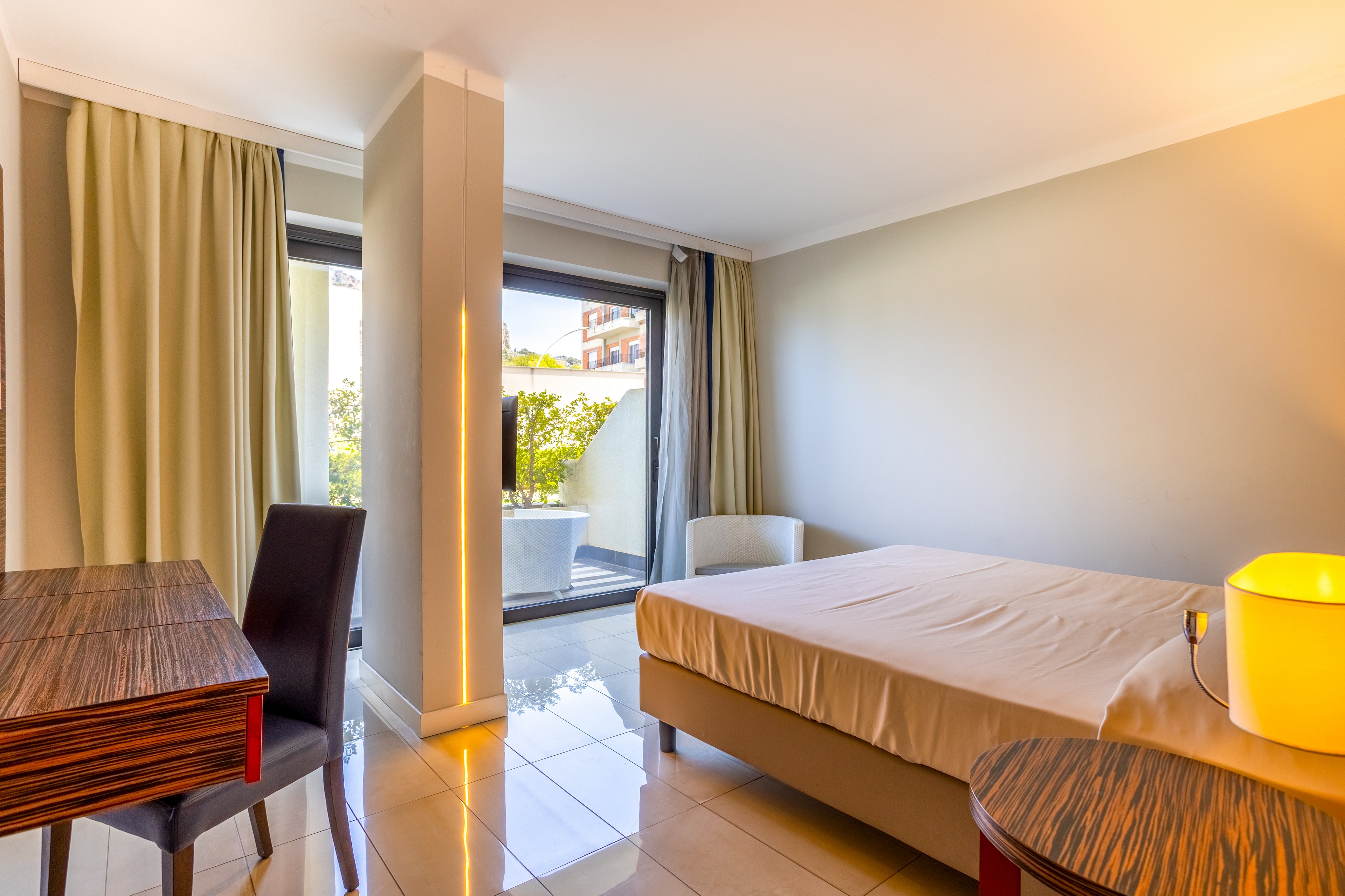 A contemporary hotel room features a neatly made bed and tiled floor in warm neutral tones. Large glass doors open to a small terrace with greenery, allowing natural light to fill the space. A wooden desk and chair sit to one side, while a round side table with a glowing lamp adds a cozy touch. Floor-to-ceiling curtains frame the window area, creating a calm and inviting atmosphere.