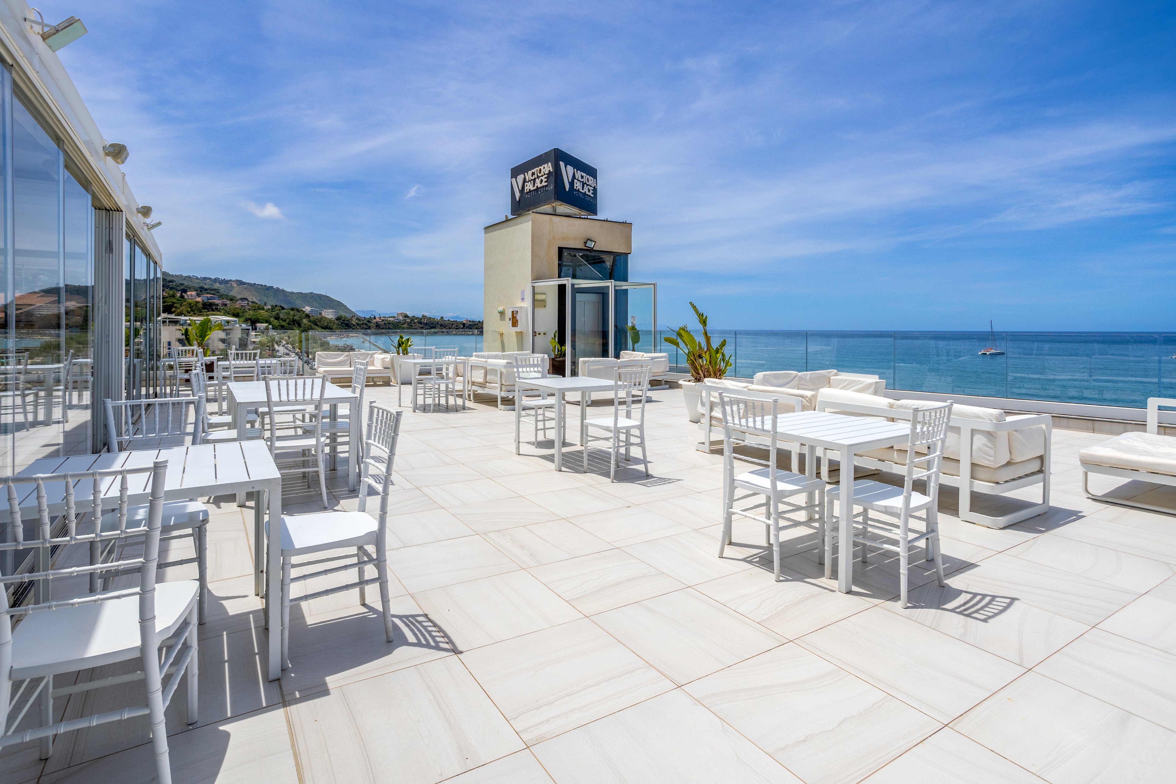 An open-air rooftop terrace features white tables, chairs, and lounge seating under a bright blue sky. Glass railings frame expansive views of the calm sea and distant coastline. A small tower structure with visible text stands near the center, while potted plants add a touch of greenery. The scene feels modern, spacious, and inviting for outdoor dining or relaxation.