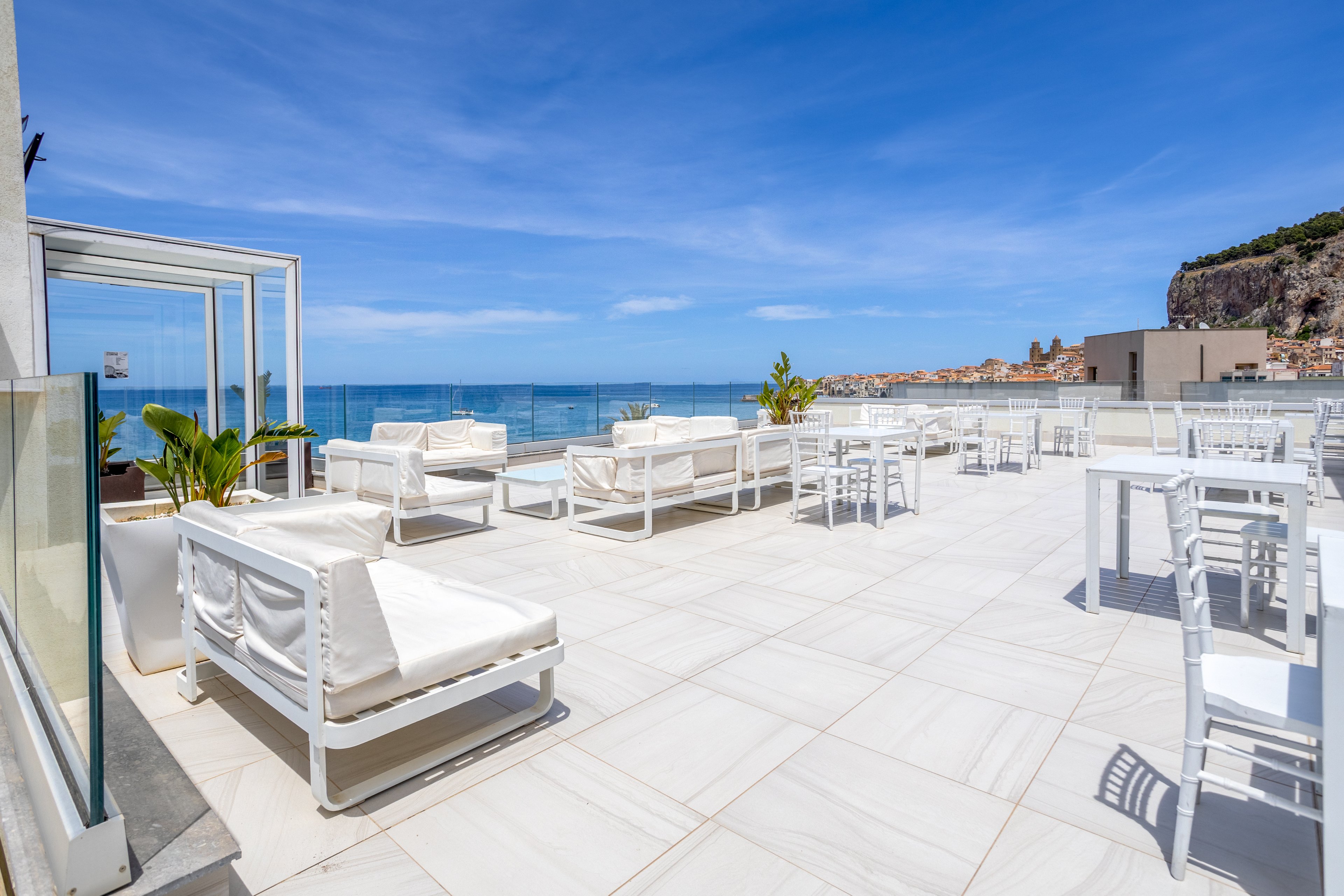 A bright rooftop terrace features white lounge sofas and dining tables under a clear blue sky. The space opens toward a calm sea with coastal buildings visible in the distance. Minimalist white furnishings and tiled flooring create a clean, modern look. Potted green plants add a touch of nature to the relaxing seaside setting.
