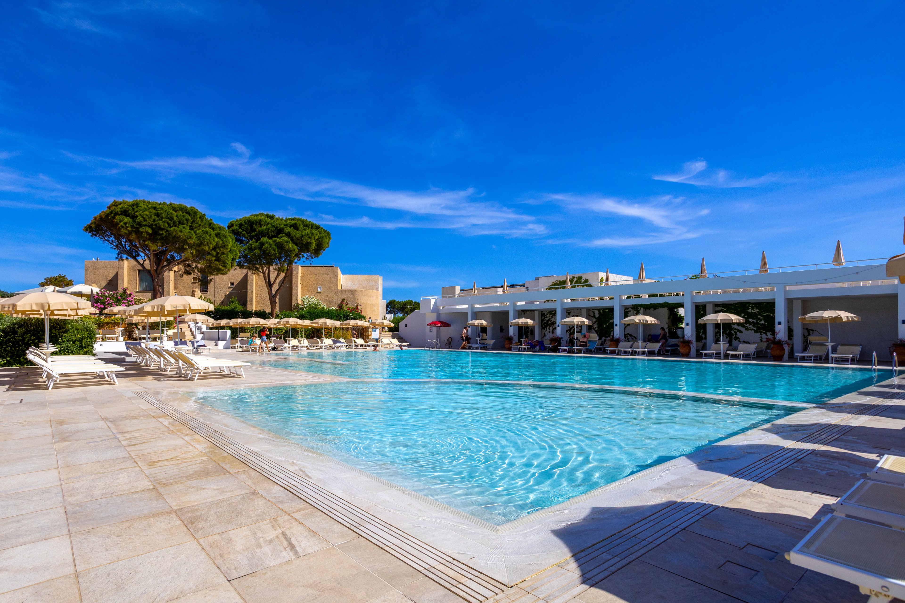 Large outdoor swimming pool at a resort is surrounded by sun loungers and umbrellas under a clear blue sky. A white poolside terrace leads to a row of cabanas or shaded seating areas. Tall trees and modern buildings frame the background, creating a relaxed vacation atmosphere. The bright sunlight highlights the turquoise water and clean architectural lines.