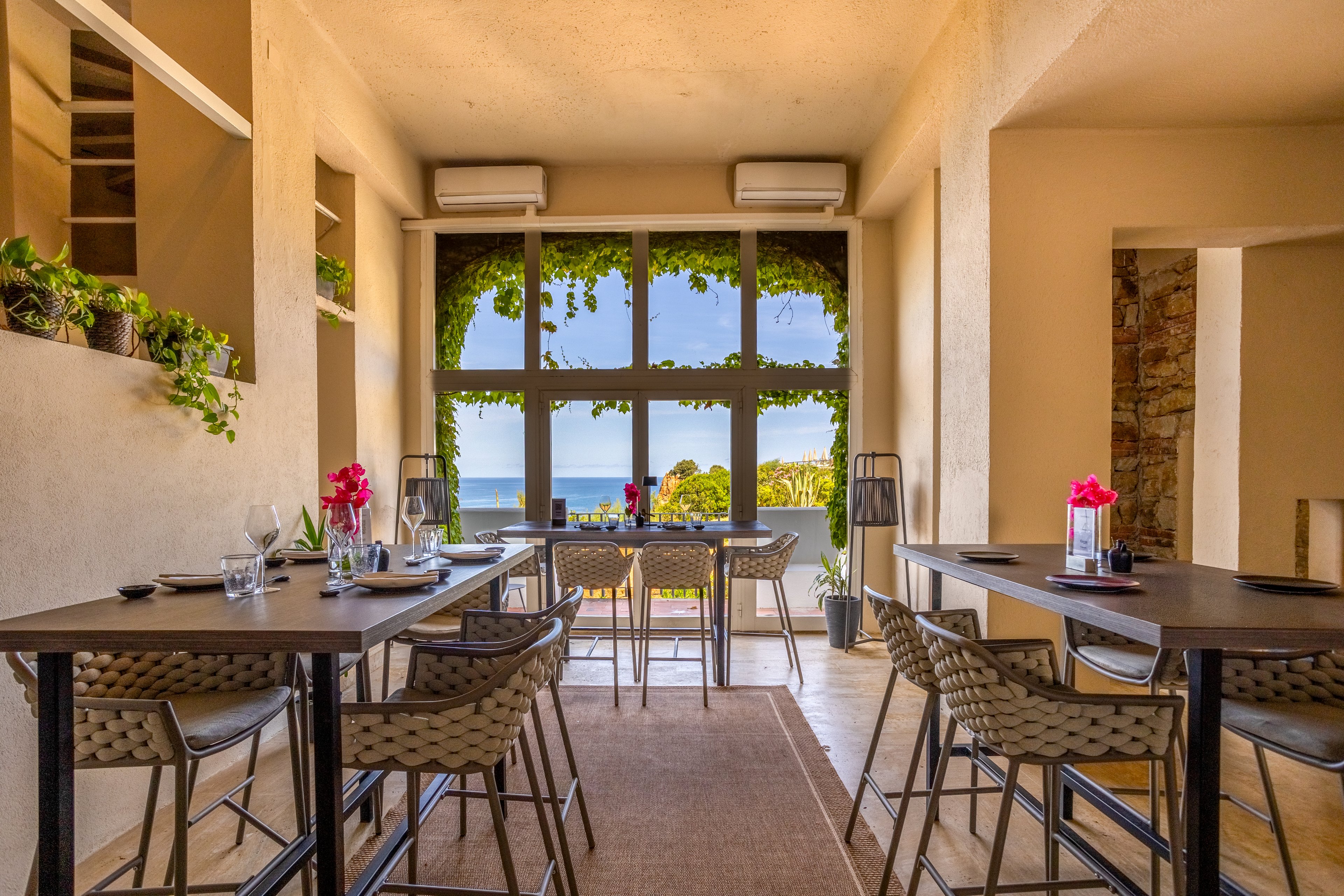 Bright restaurant interior features tall tables and woven bar stools arranged along a narrow dining space. Large windows at the far end frame a scenic view of the ocean and palm trees outside. Green vines hang around the window, adding a natural touch to the warm beige walls. Table settings with glasses, plates, and pink flowers create a relaxed yet elegant atmosphere.