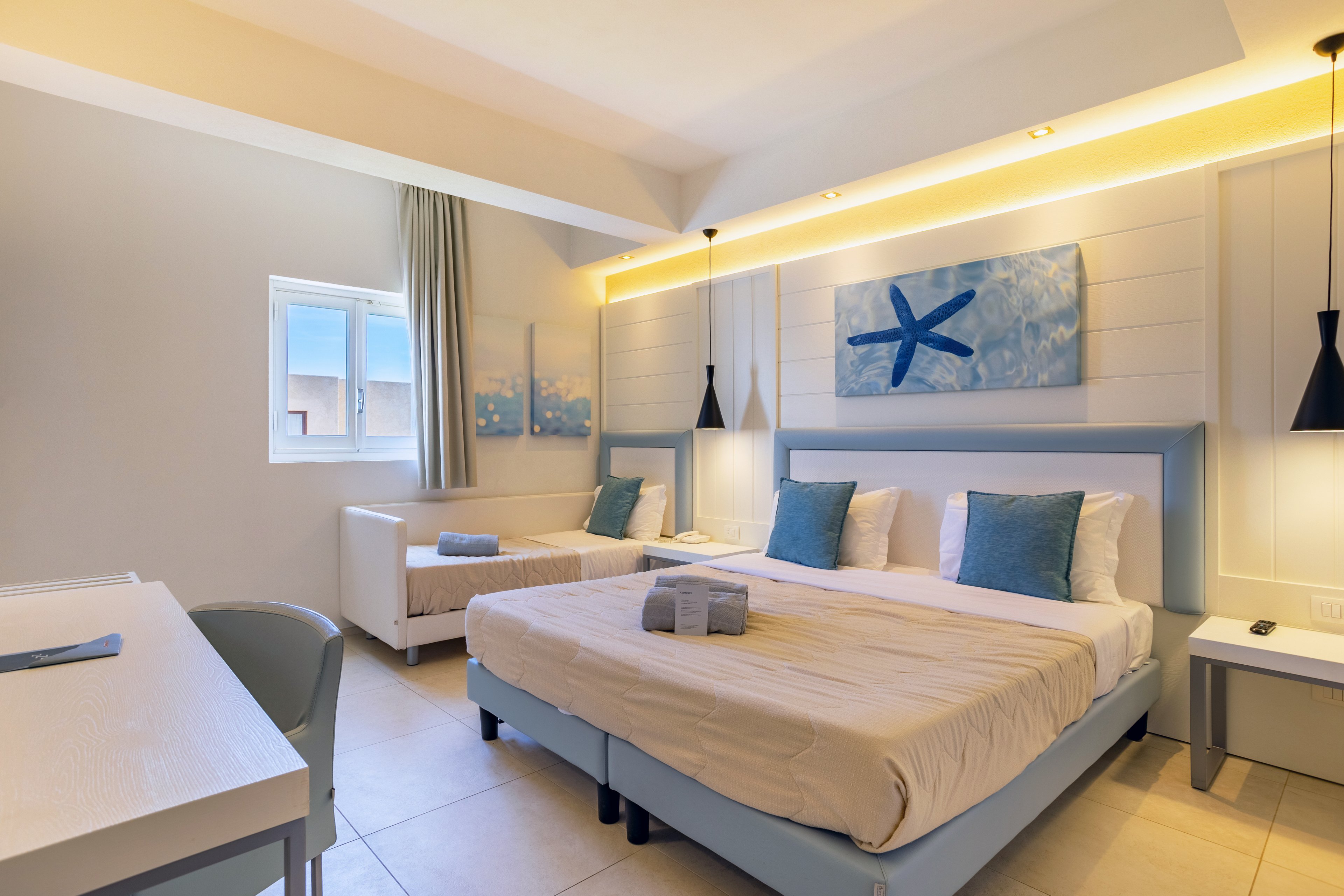Contemporary hotel room featuring a large double bed and a smaller single bed with beige linens and blue accent pillows. The space is decorated in a coastal style with starfish artwork above the headboard and soft ambient lighting along the wall. A small desk and chair sit in the foreground, while a window with curtains brings in natural light. Rolled towels are neatly placed on the main bed, enhancing the clean and welcoming atmosphere.