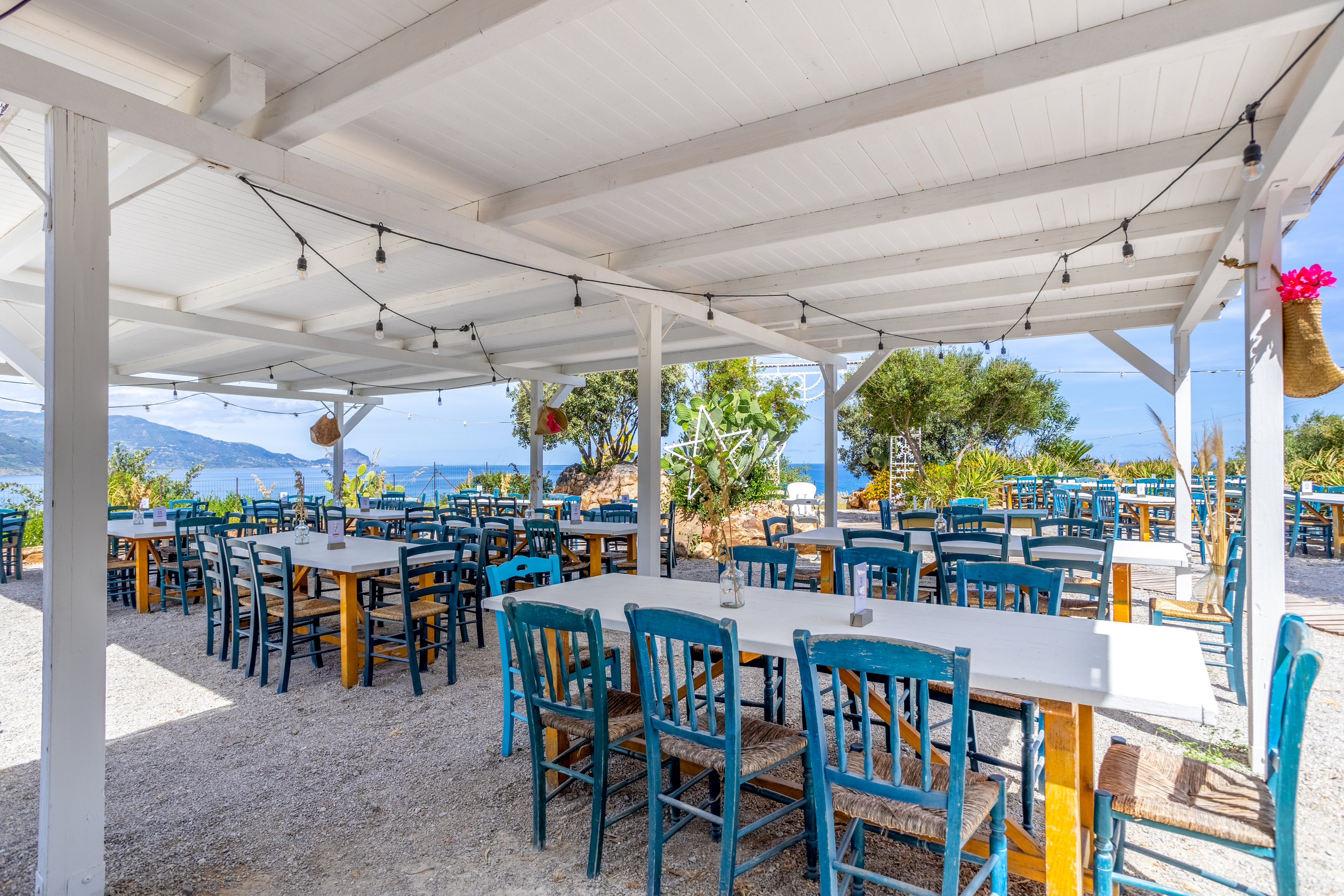 An open-air restaurant with white tables and bright blue wooden chairs sits under a white pergola. The dining area overlooks a scenic coastline with blue water and distant hills. String lights hang from the ceiling, and potted plants and trees add greenery around the seating. The atmosphere feels relaxed and inviting, ideal for casual outdoor dining by the sea.