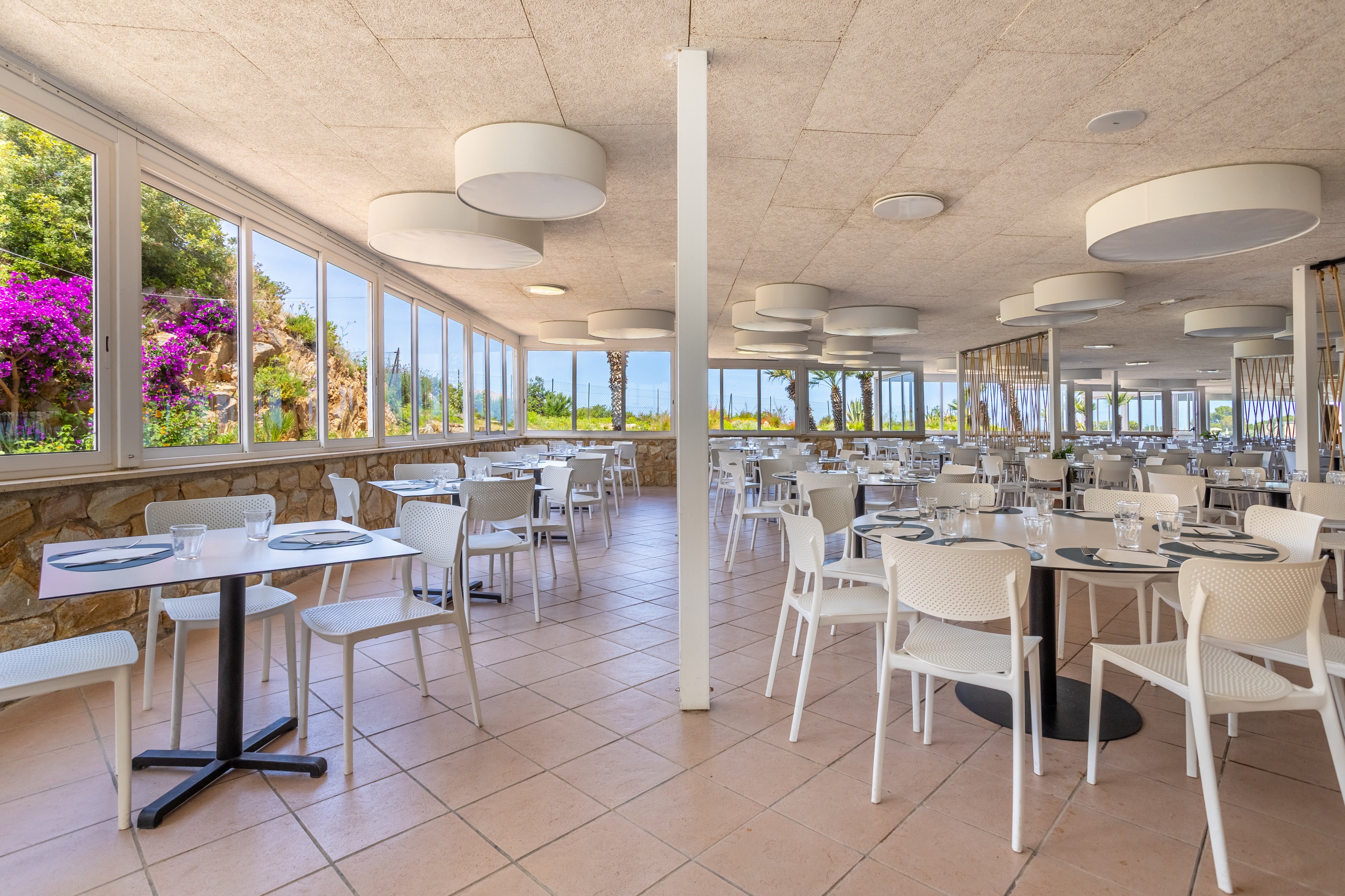 Spacious restaurant interior featuring rows of white tables and chairs set for dining. Large windows line the walls, offering views of lush greenery and vibrant purple flowers outside. The room is filled with natural light, complemented by round ceiling fixtures and tiled flooring. Table settings include plates, glasses, and cutlery, creating a clean, contemporary atmosphere ready for guests.