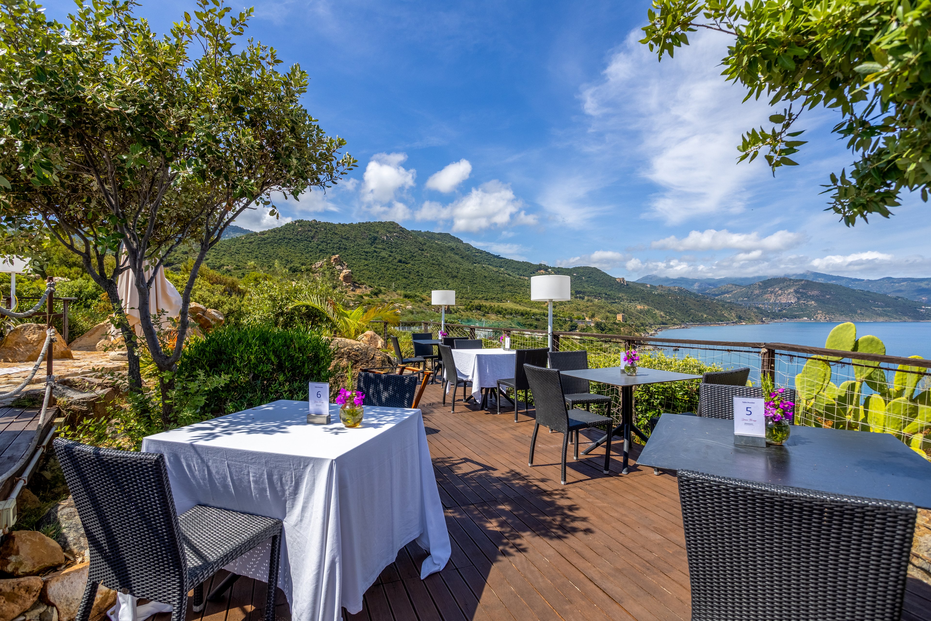 An outdoor restaurant terrace is set with tables and wicker chairs on a wooden deck. White tablecloths, small flower arrangements, and numbered table cards create a casual dining atmosphere. The terrace is surrounded by lush greenery, trees, and coastal plants, with a railing framing the view. In the background, rolling hills and a blue sea stretch under a bright, partly cloudy sky.
