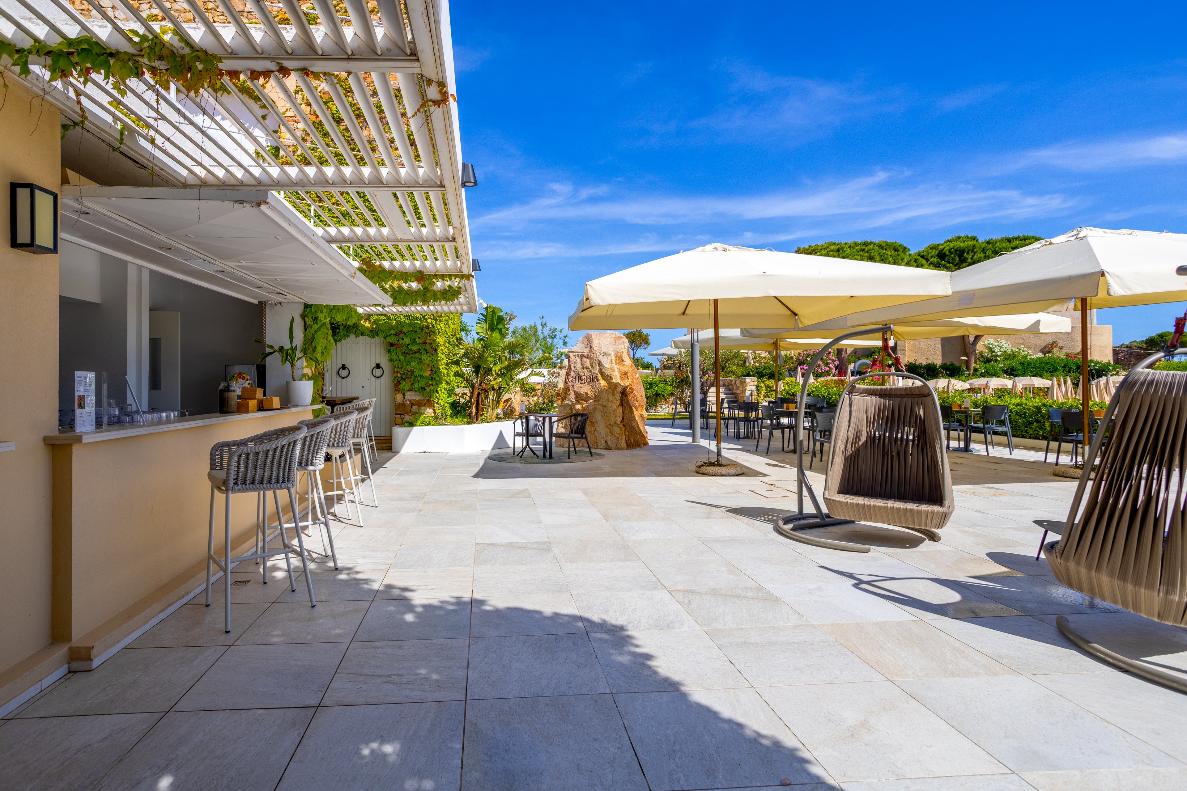 An open-air bar terrace features modern stools and a clean counter under a white pergola. Swing chairs and large cream-colored umbrellas provide shaded seating on the spacious tiled patio. Lush greenery and a bright blue sky create a relaxed vacation atmosphere. The scene suggests a tranquil resort or hotel outdoor lounge area.