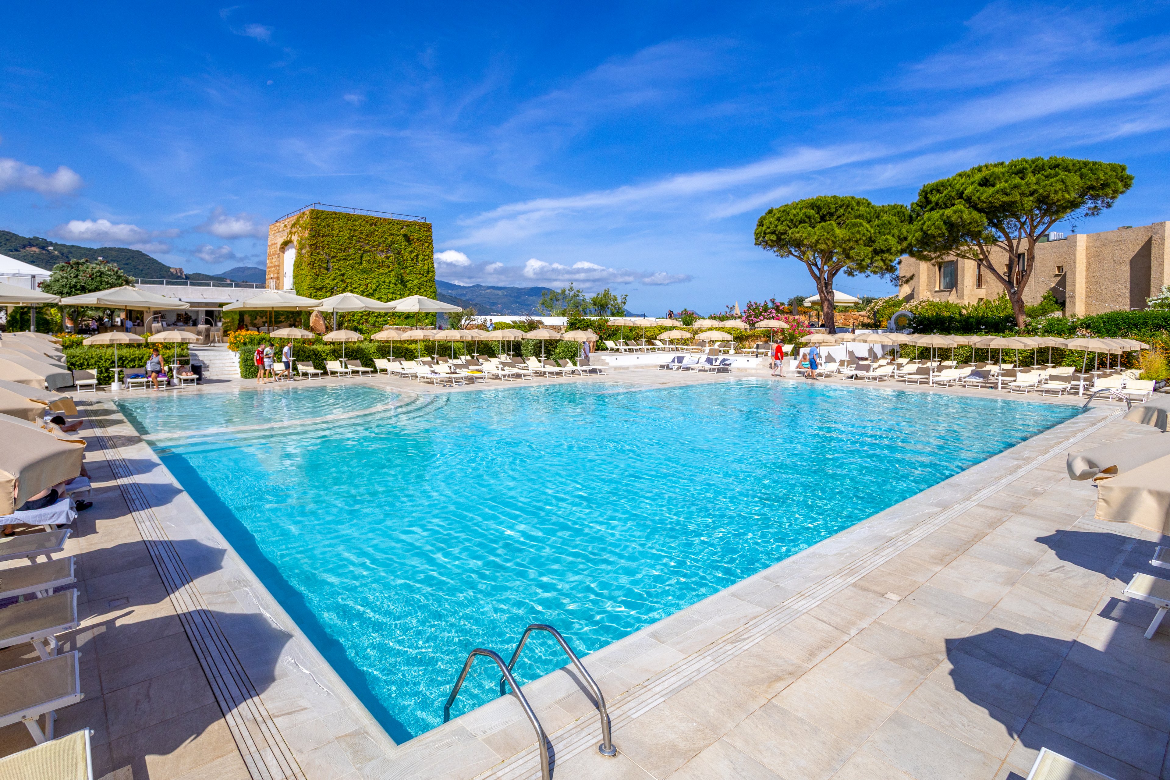 Large turquoise swimming pool at a resort is surrounded by white sun loungers and umbrellas under a bright blue sky. Guests relax around the poolside area, with landscaped greenery and distinctive trees framing the scene. A building covered in ivy and other low-rise structures create a Mediterranean-style backdrop. The overall atmosphere is calm, inviting, and ideal for a summer holiday.