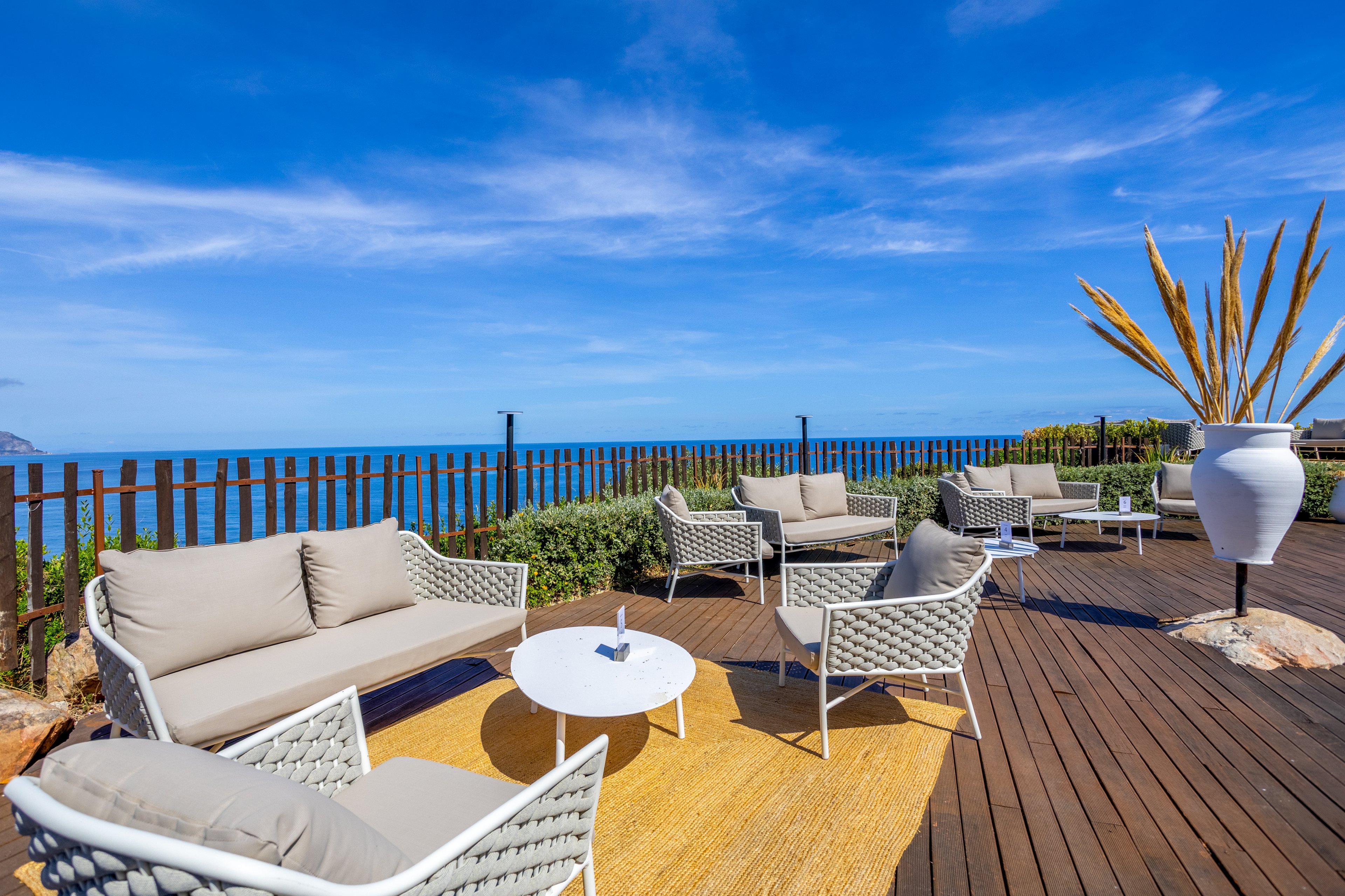 An outdoor terrace features modern wicker lounge chairs with beige cushions arranged around small white tables. The space is set on a wooden deck overlooking a bright blue ocean under a clear sky. A large white decorative planter with tall dried plants adds a sculptural element to the scene. The overall atmosphere is relaxed and inviting, ideal for seaside leisure and socializing.