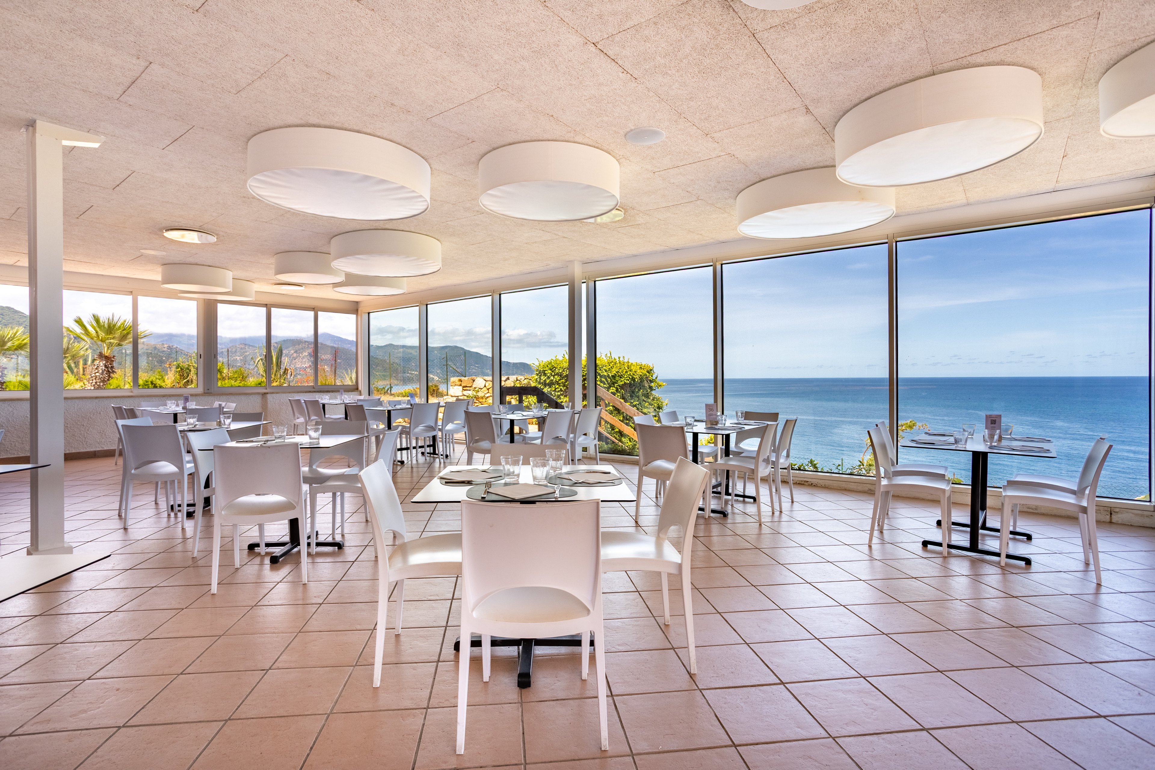 Spacious modern restaurant interior features white tables and chairs arranged across a tiled floor. Large floor-to-ceiling windows reveal a scenic ocean view with distant mountains and coastal greenery. Circular ceiling lights add a contemporary touch and enhance the bright, airy atmosphere. The setting appears calm and inviting, ideal for relaxed seaside dining.