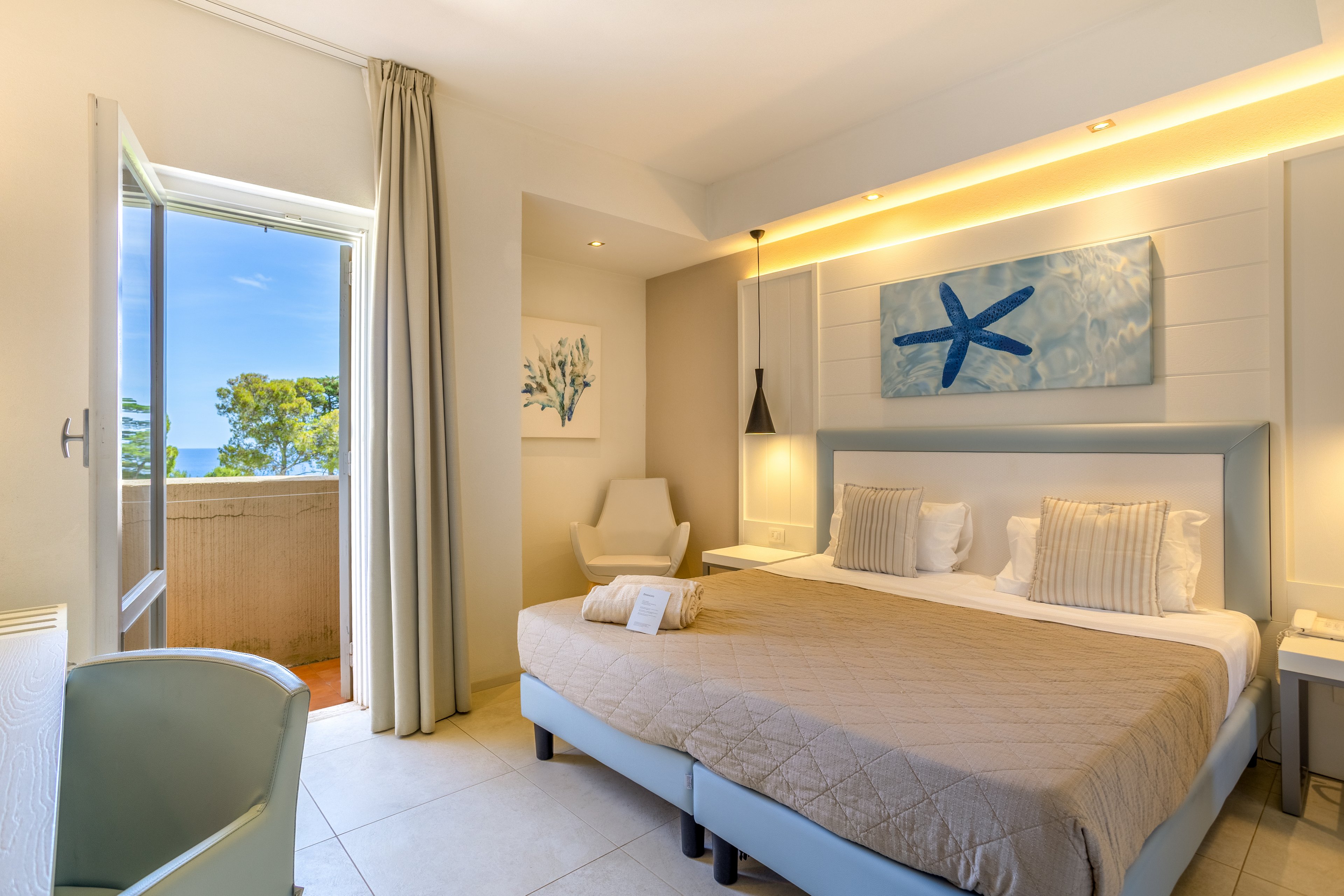 A modern hotel bedroom features a large double bed with beige bedding and multiple pillows. The room is decorated in a coastal style, including a starfish artwork above the bed and a framed sea-themed painting on the wall. Natural light streams in from an open balcony door, revealing a glimpse of blue sky and greenery outside. Soft neutral tones, minimalist furniture, and subtle accent lighting create a calm and inviting atmosphere.